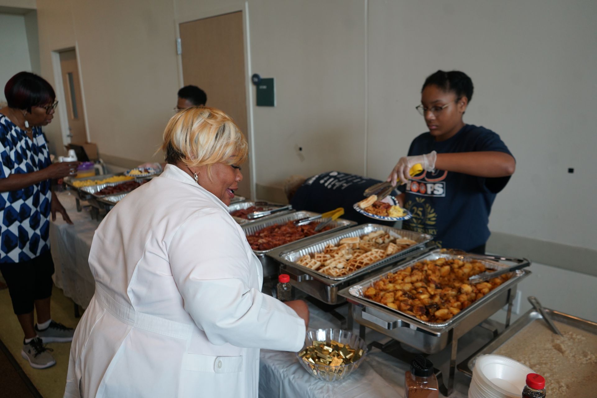 People serving food from buffet: Various dishes in chafing dishes, one person in white coat.