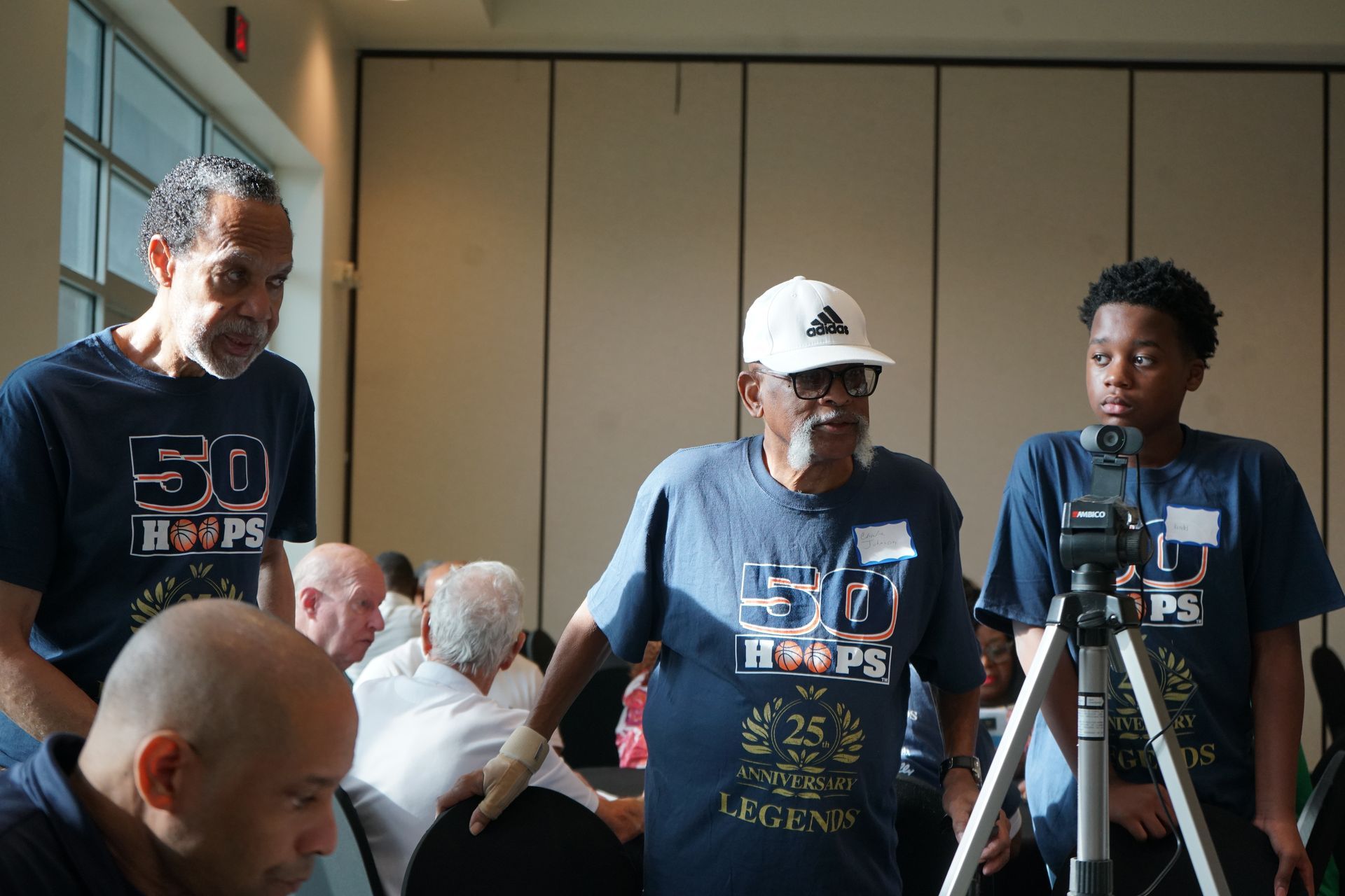 Three men in matching blue t-shirts; one with a hat and microphone, standing indoors. Other people are seated.