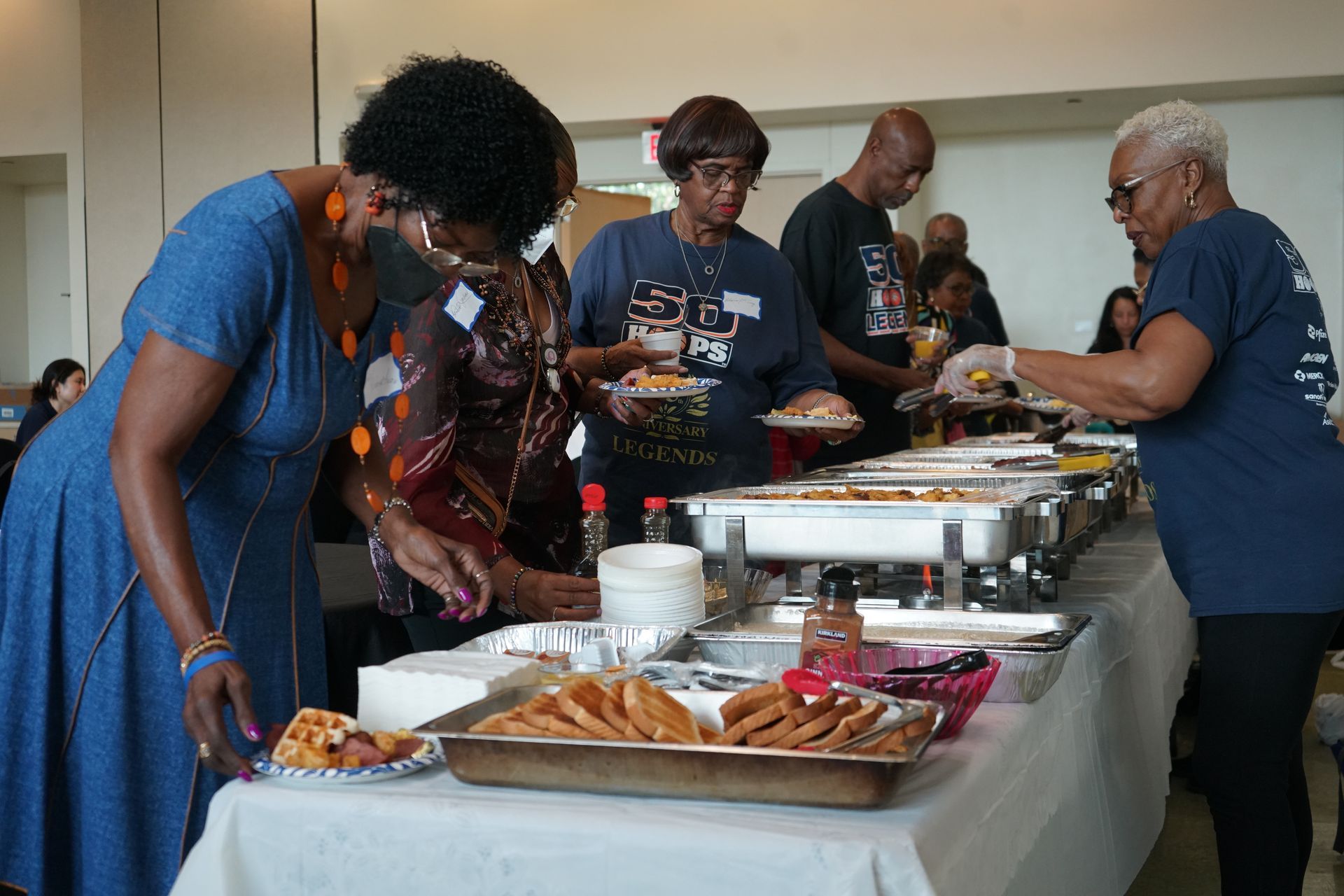 People at a buffet table, serving food from warming trays. Sunlight floods the room.