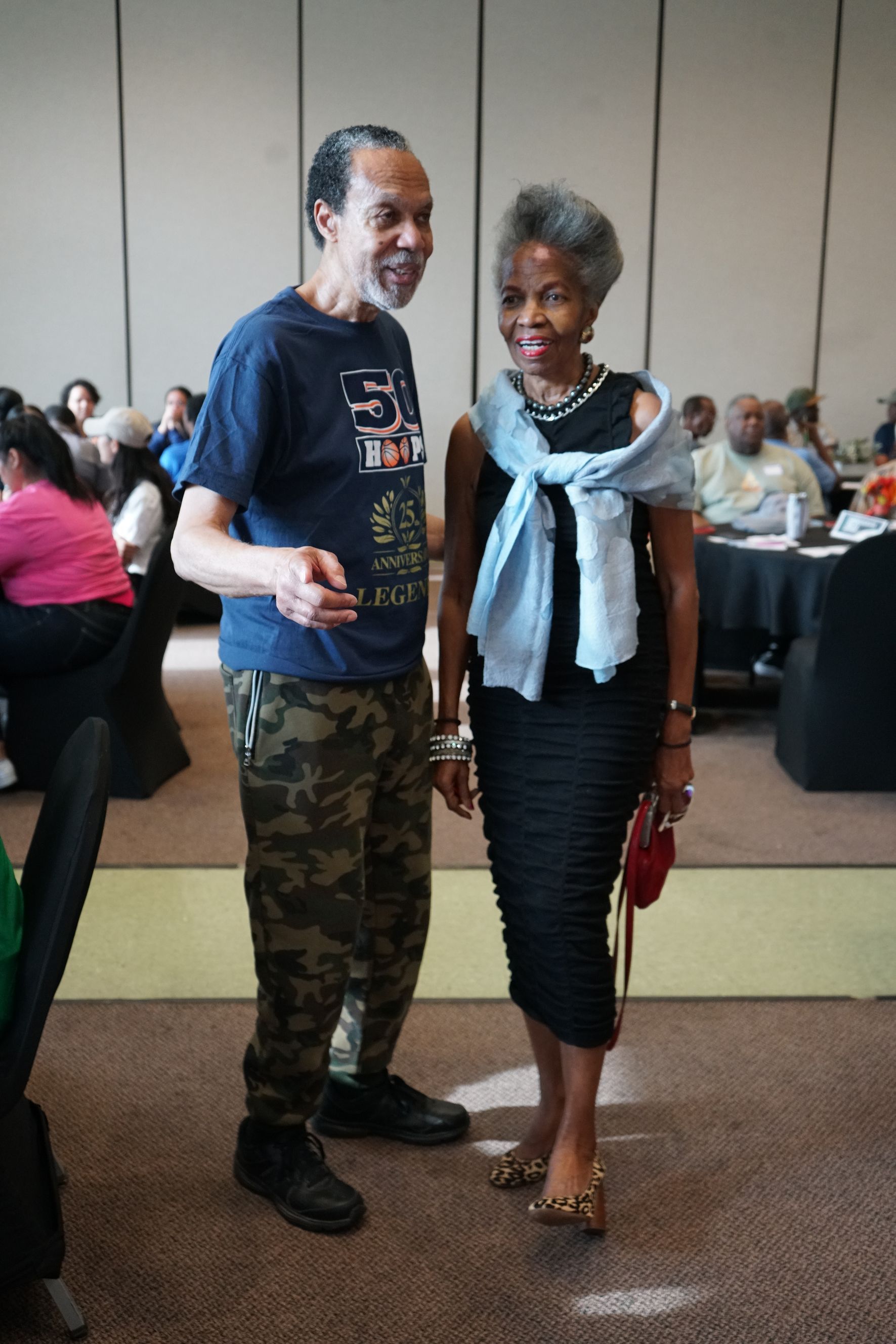 Man in camouflage pants and woman in black dress pose indoors.