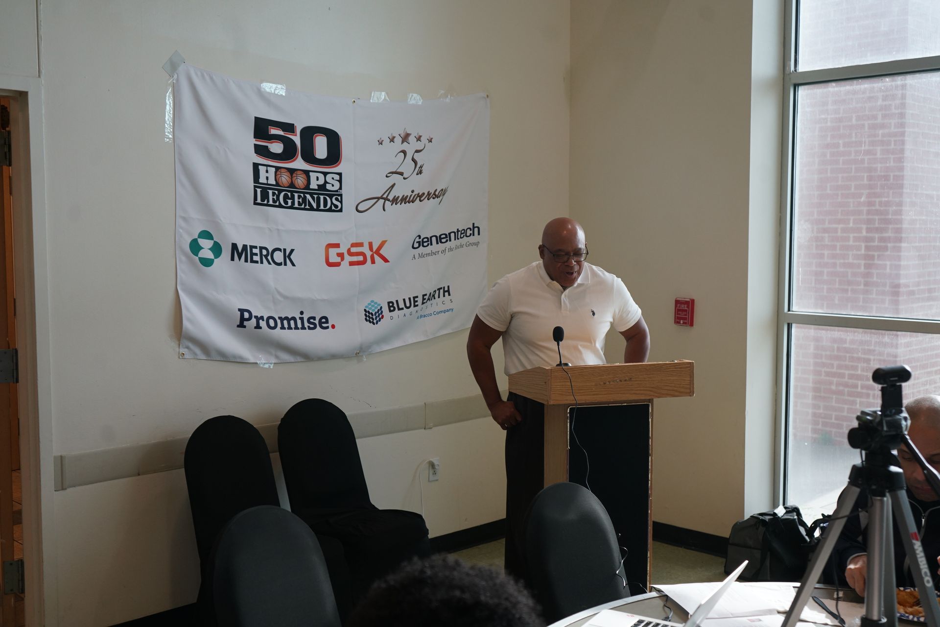 Man speaking at a podium in front of a banner, near a window.  Chairs are to the left, and a tripod is to the right.