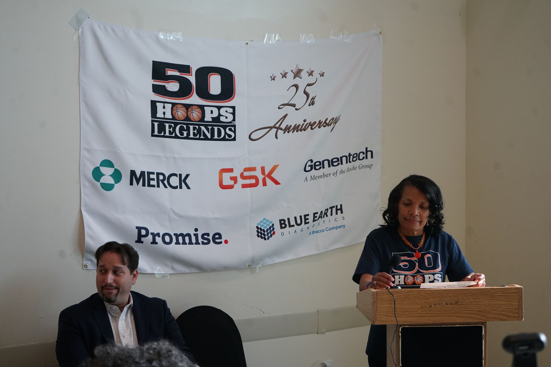Woman speaking at podium, banner with logos behind her. Man sits to the left.
