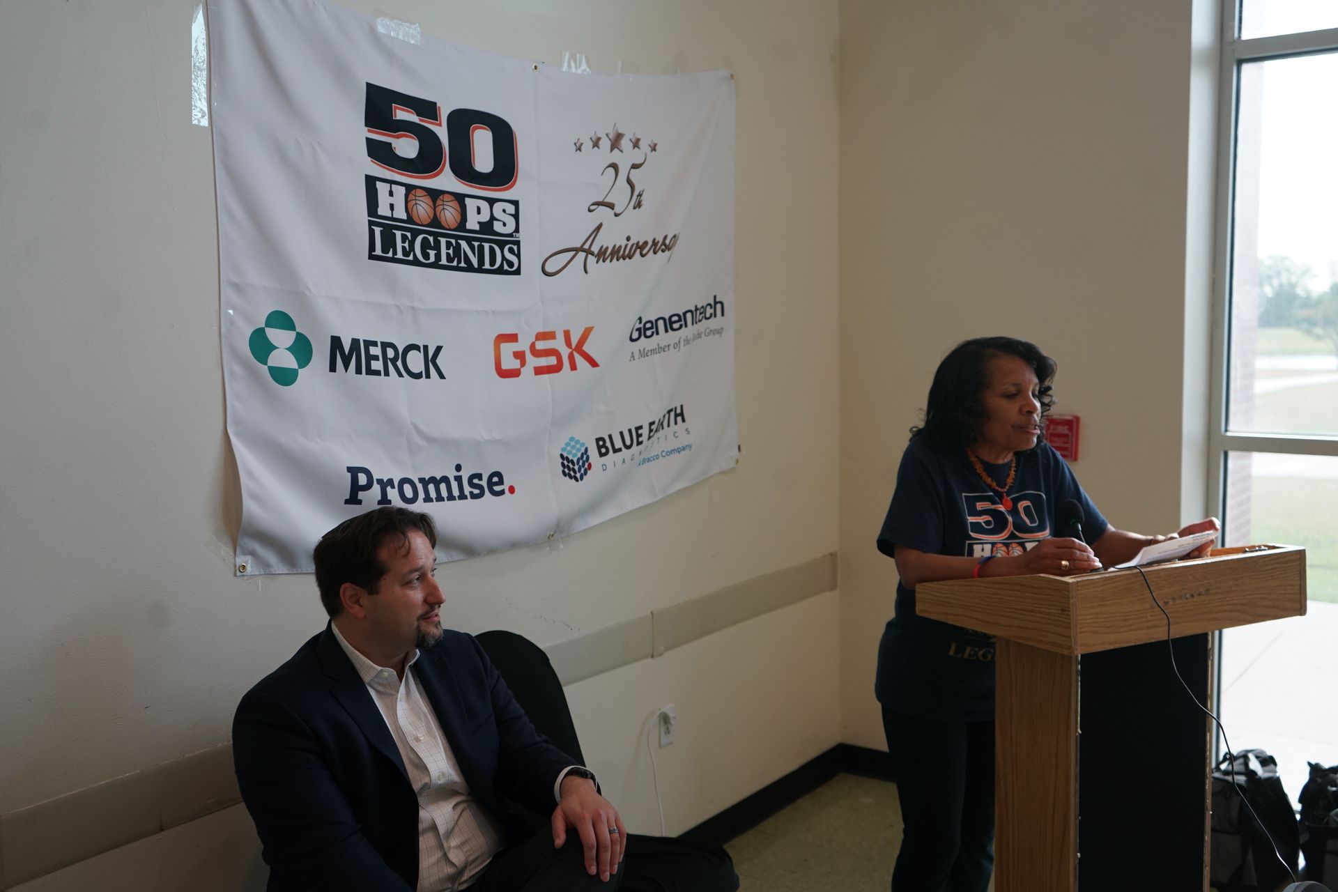 Woman speaking at a podium; banner with sponsors and 