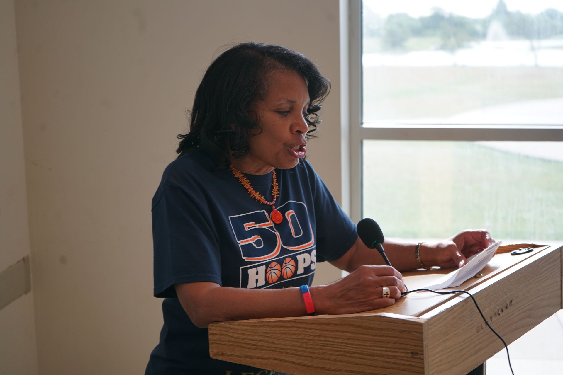 Woman speaking at a podium, reading from paper. She wears a blue shirt and orange necklace; a window is behind her.
