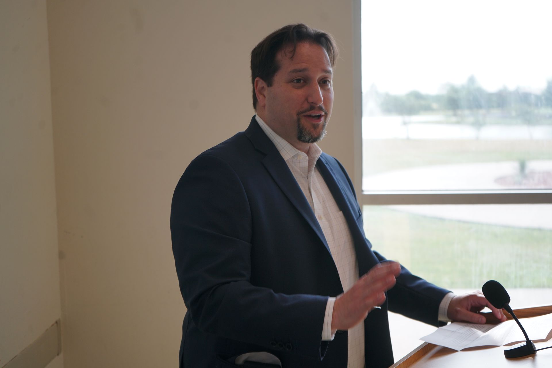 Man in suit speaking at a podium, gesturing with his hands. Window and papers visible.