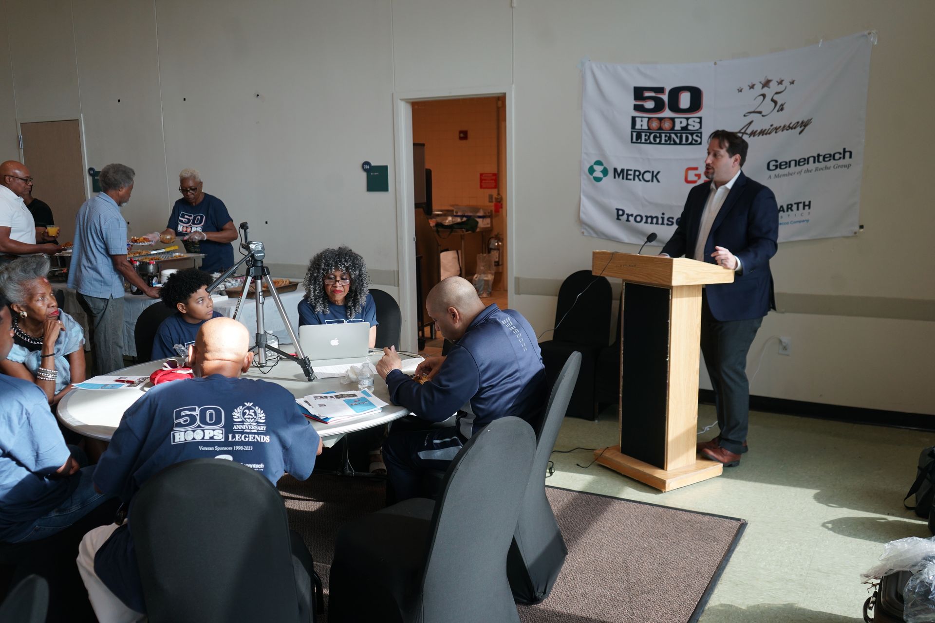 Man speaking at a podium; people seated at round tables in a room with a 