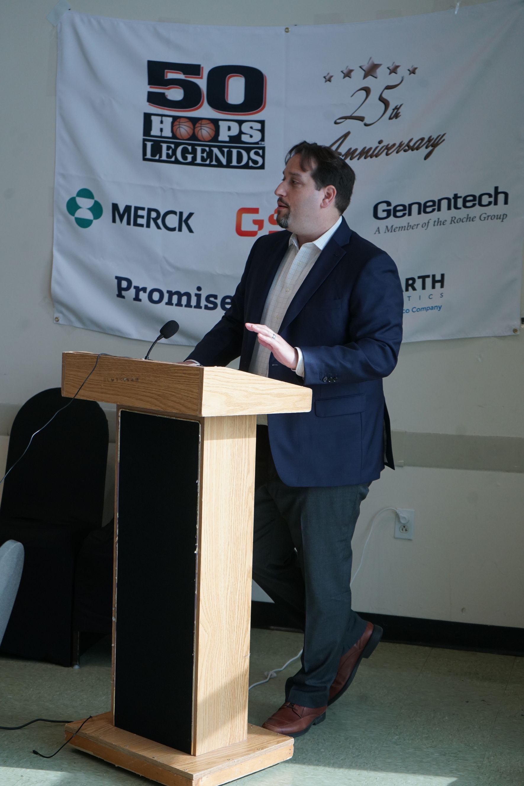 Man in navy blazer speaks at a podium; banner for 