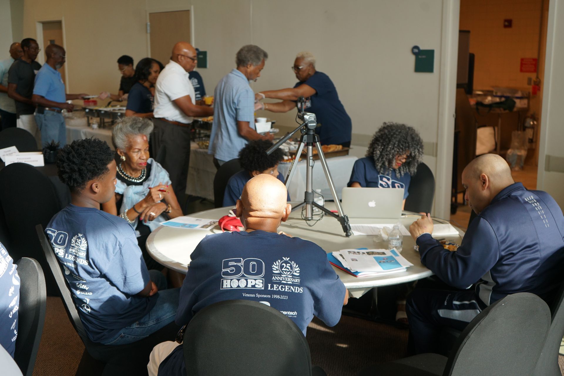 People at a table working on laptops and papers, in a brightly lit room. Some stand at a buffet.