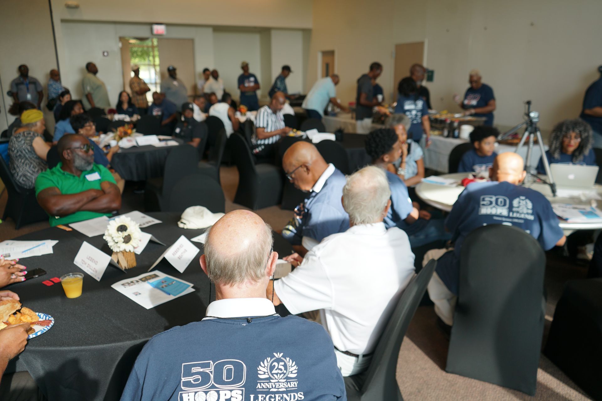 People seated at round tables in a large room, some with food and drinks. Many wear blue shirts, gathered for an event.