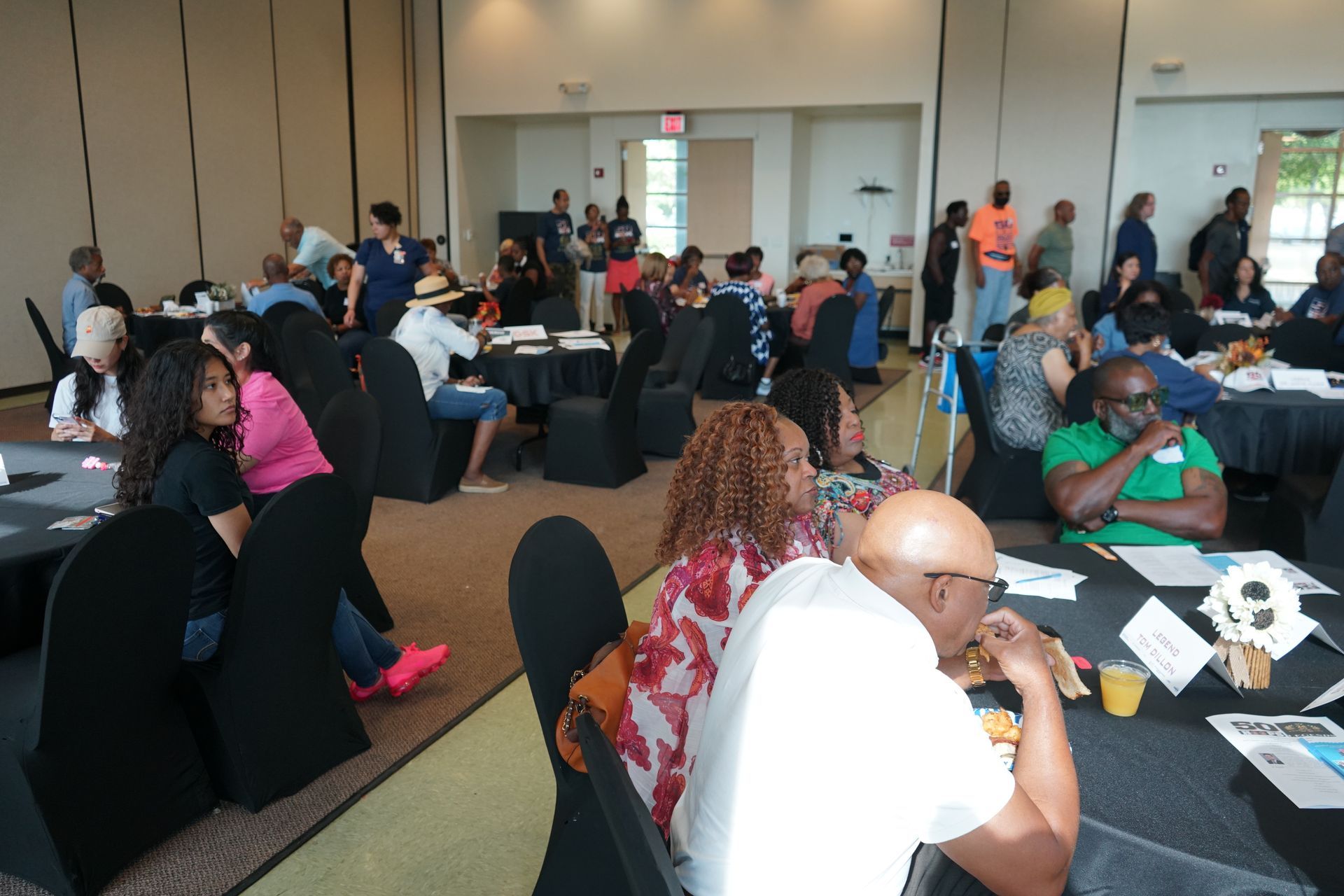 People gather around tables in a conference room. Some are talking, others are eating and drinking.