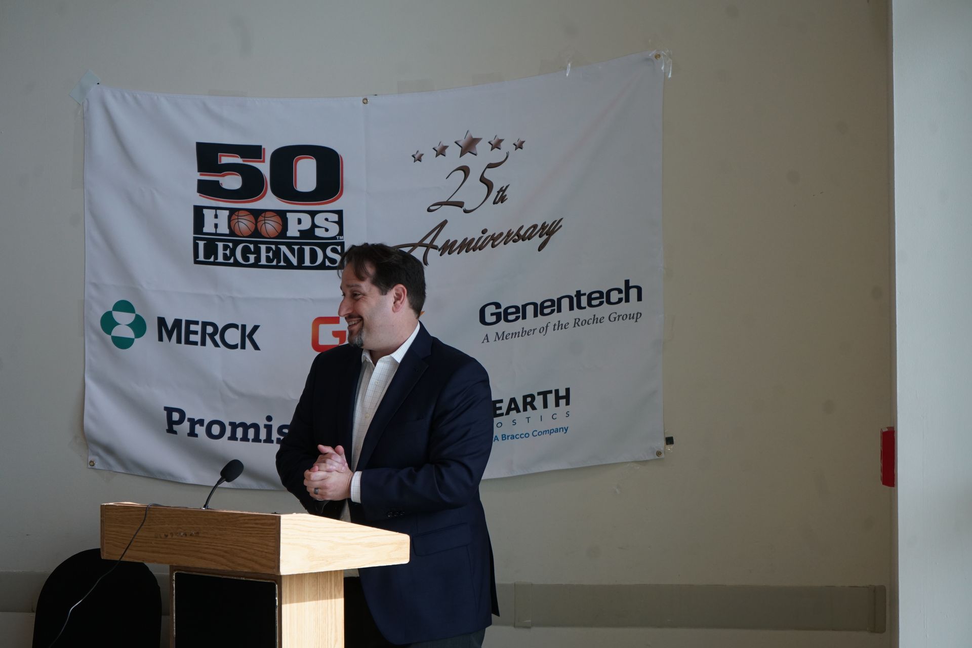 Man in suit at podium, clapping, in front of a banner for a 