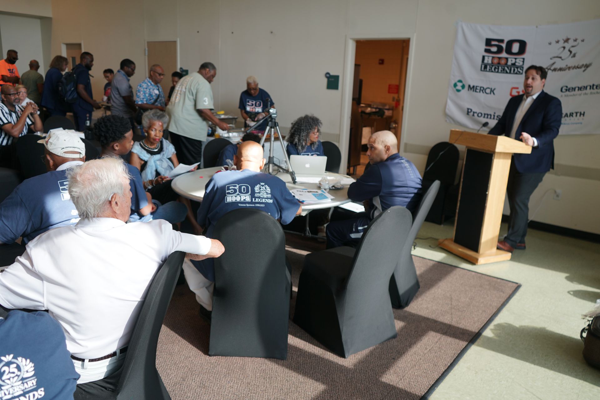 A person speaks at a podium while a group of people sit and listen in a meeting room.