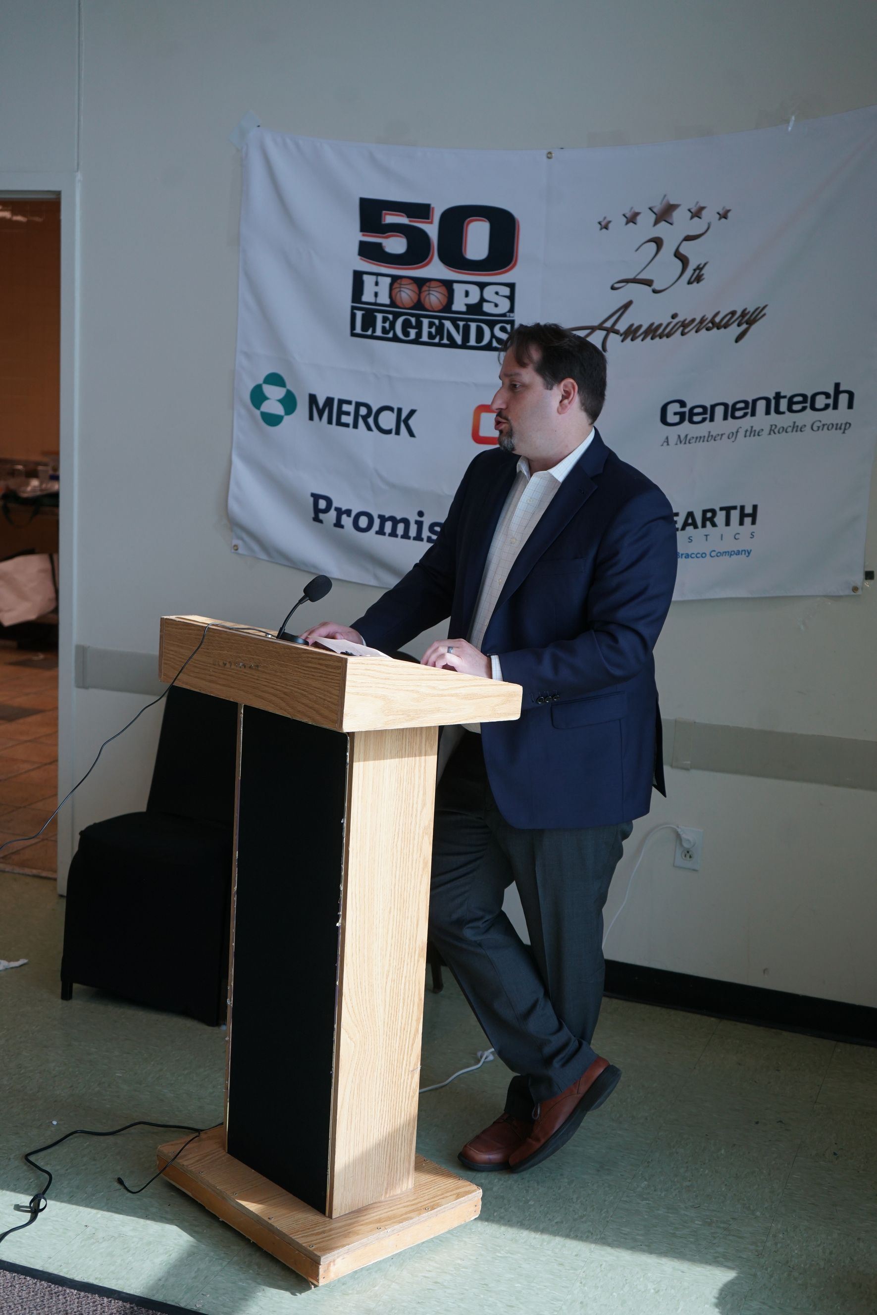 Man in blazer stands at a podium with microphone, speaking in front of a banner.