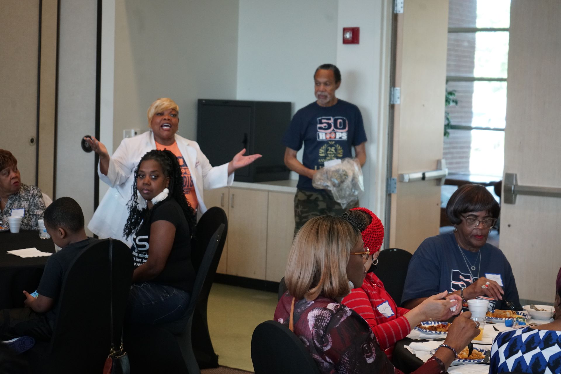 People at tables, one woman gestures, another holds items, in a room.