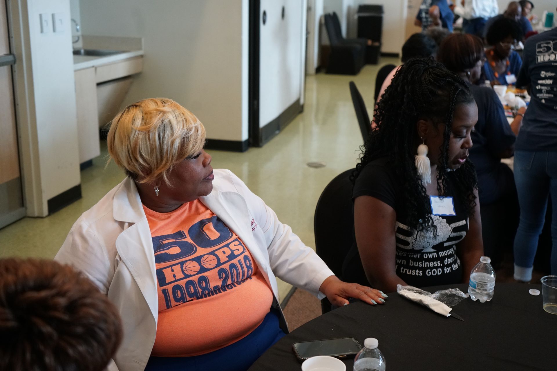 Woman in an orange shirt and blazer leans toward woman at a table, both attending an event.