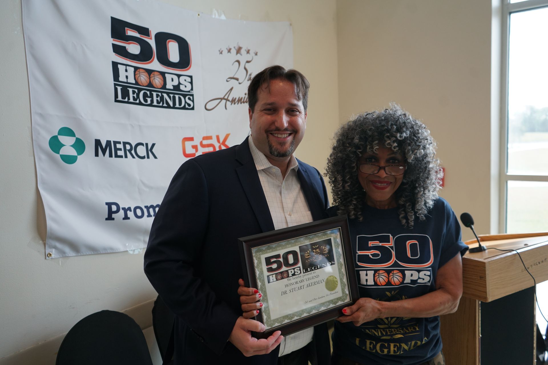 Man presents framed certificate to a woman, both holding it in front of a banner that reads 