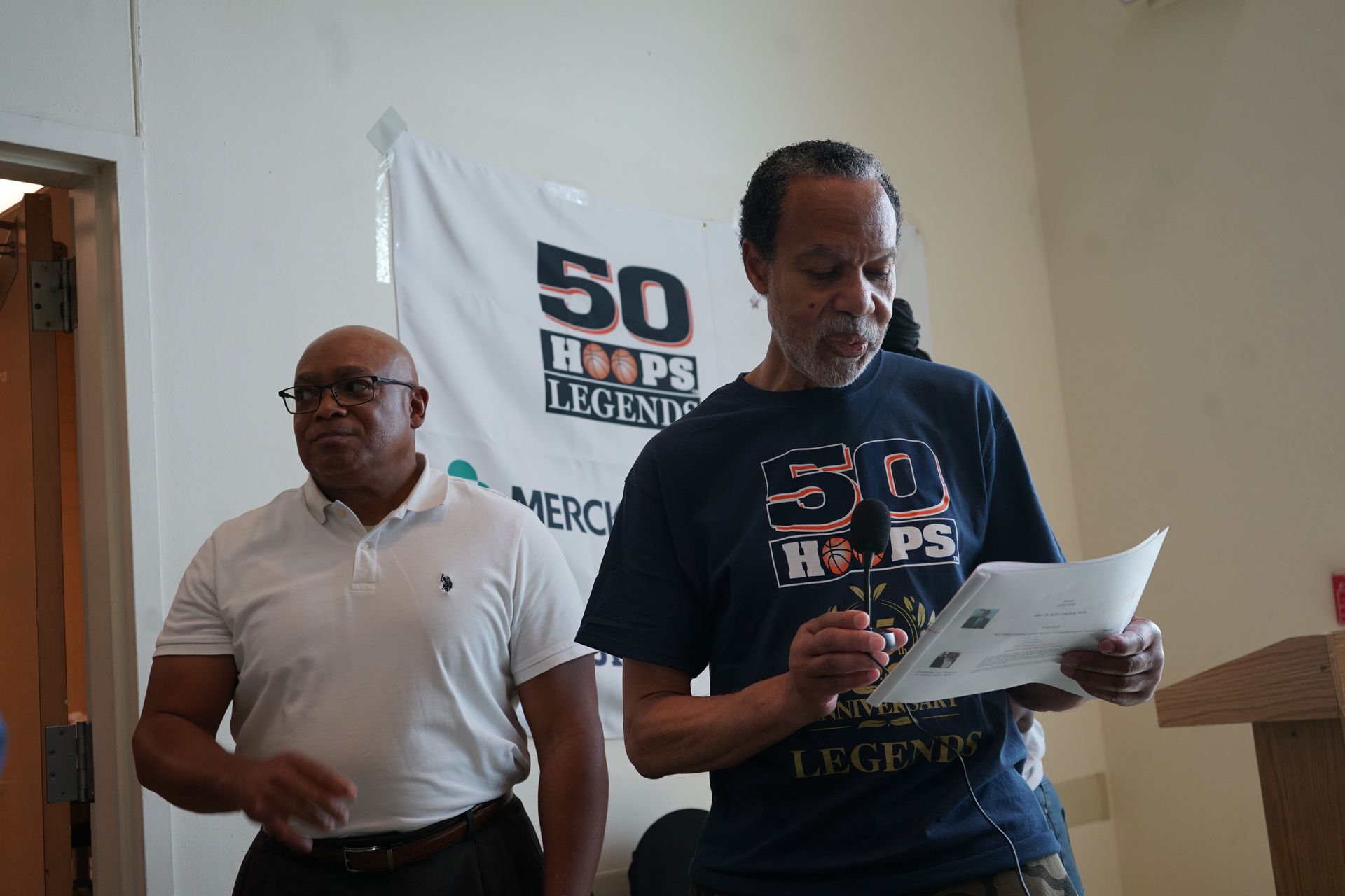 Two men standing, one reading a paper, the other looking on, in front of a banner that says 