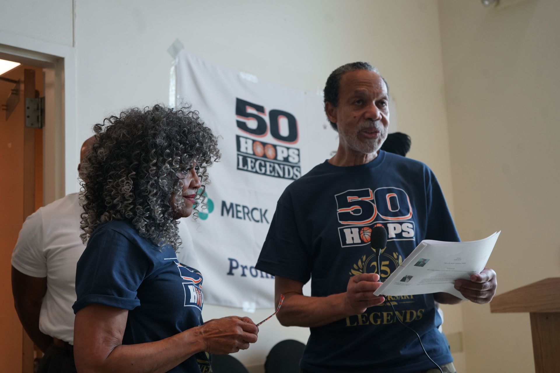 Two people stand before a banner that reads “50 Hoops Legends.” One holds a paper and mic.
