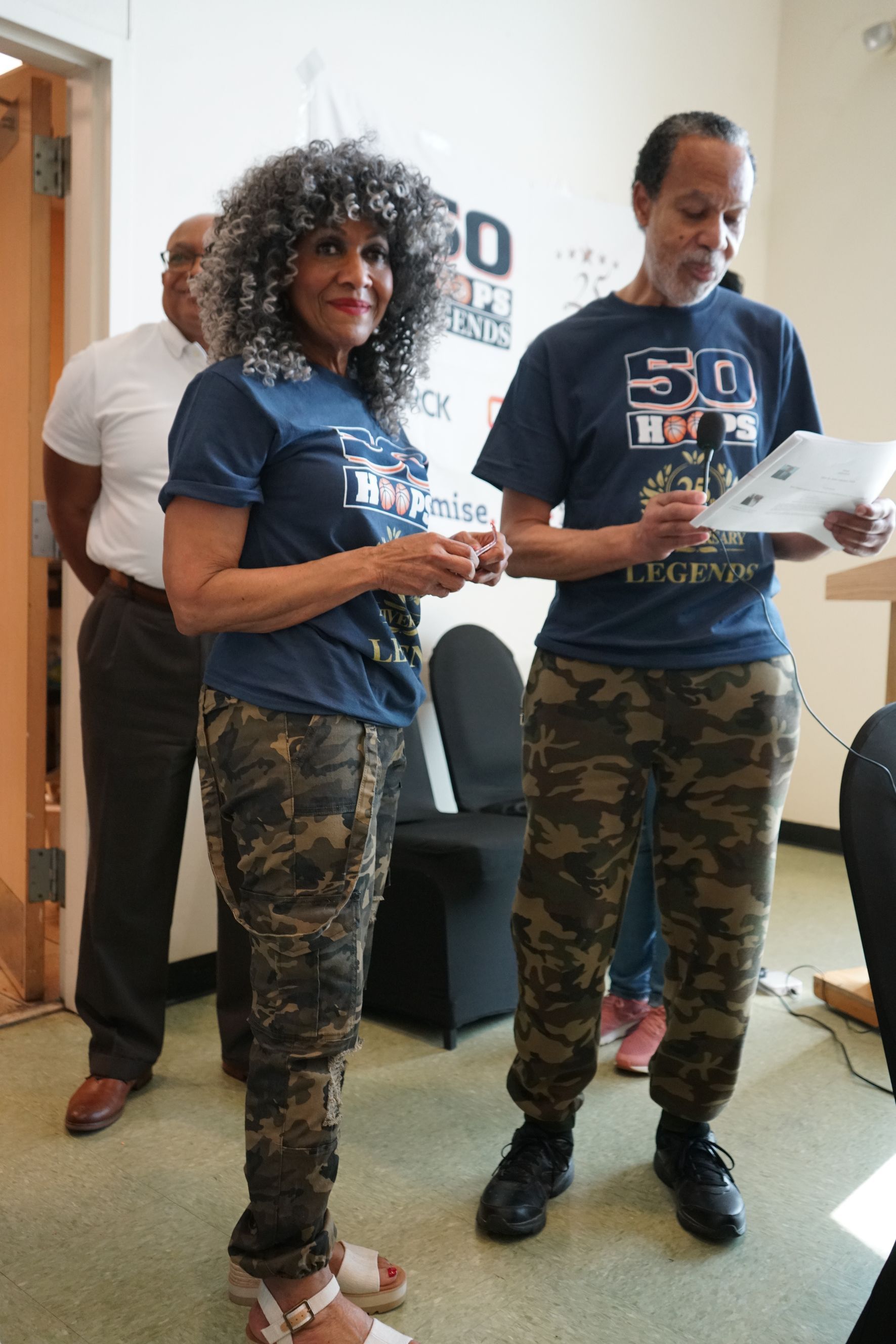 Two people in matching t-shirts and camouflage pants standing indoors, speaking at an event.