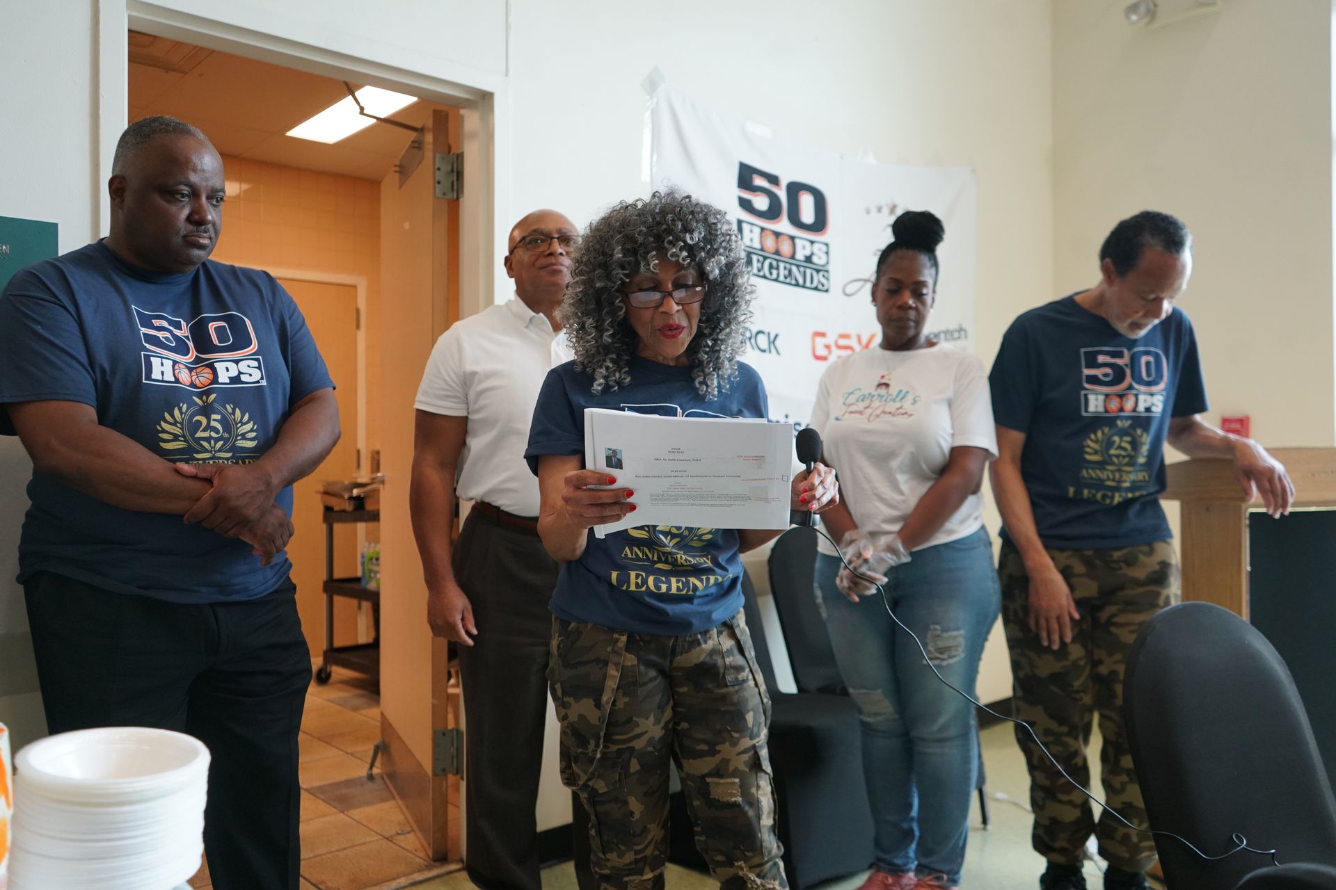 Group of people at an event; woman reads from paper, others watch; banner in background.
