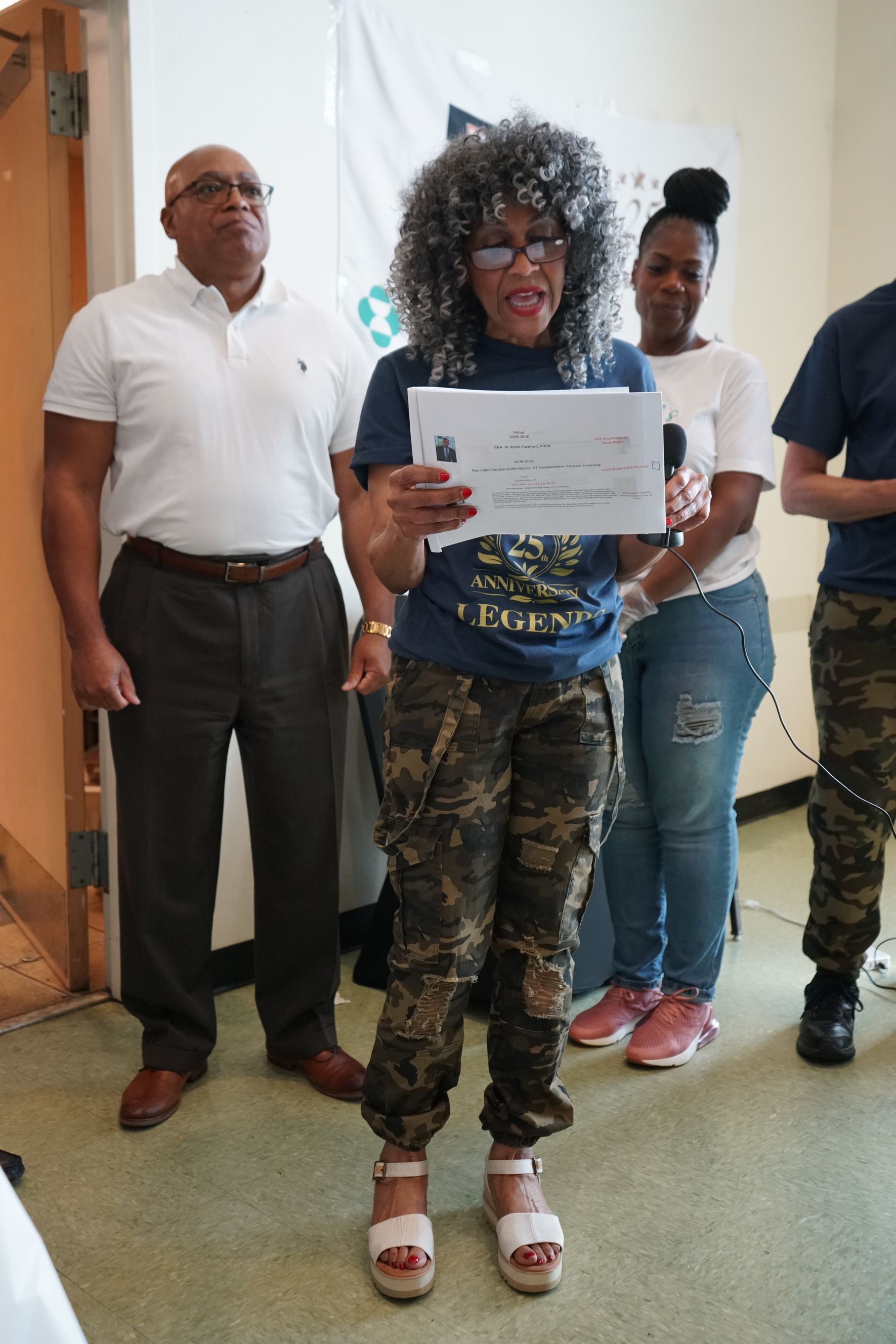 Woman in camo pants reads a document, with others behind her. Light-colored room.