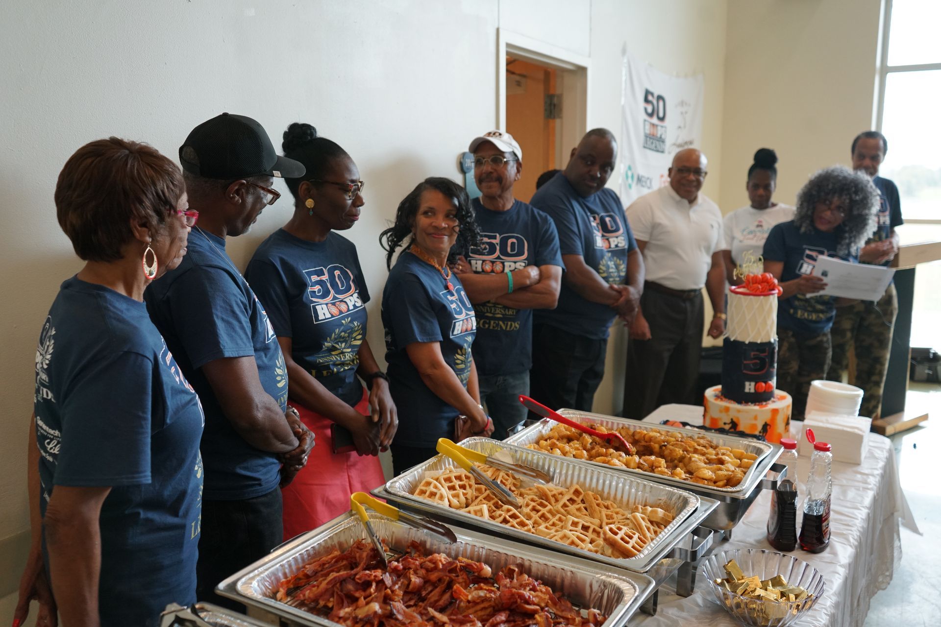 People at a party with food and a cake. Several wear blue shirts. Table set with buffet.