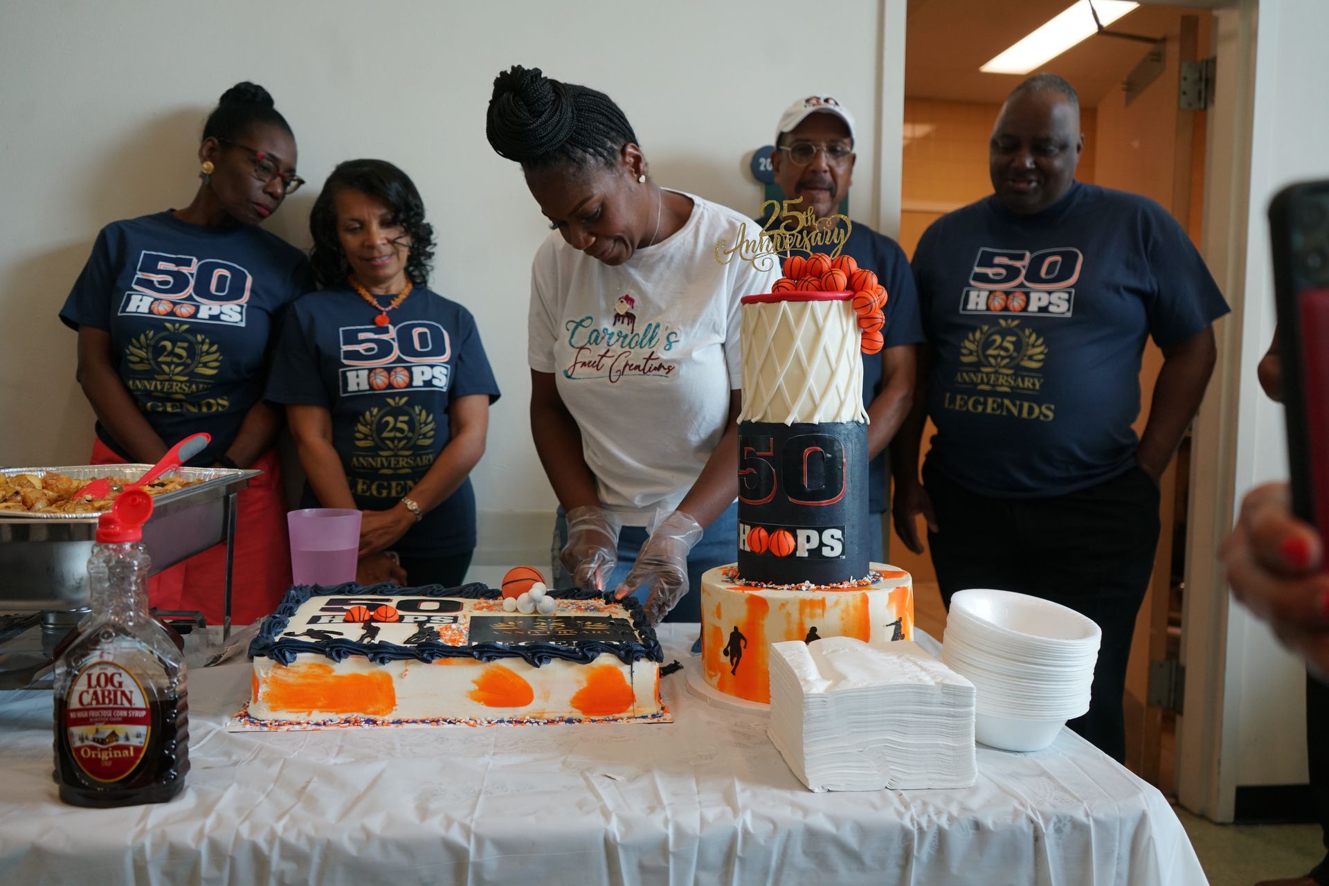 Group of people at a party, cutting a decorated cake. The cake reads 