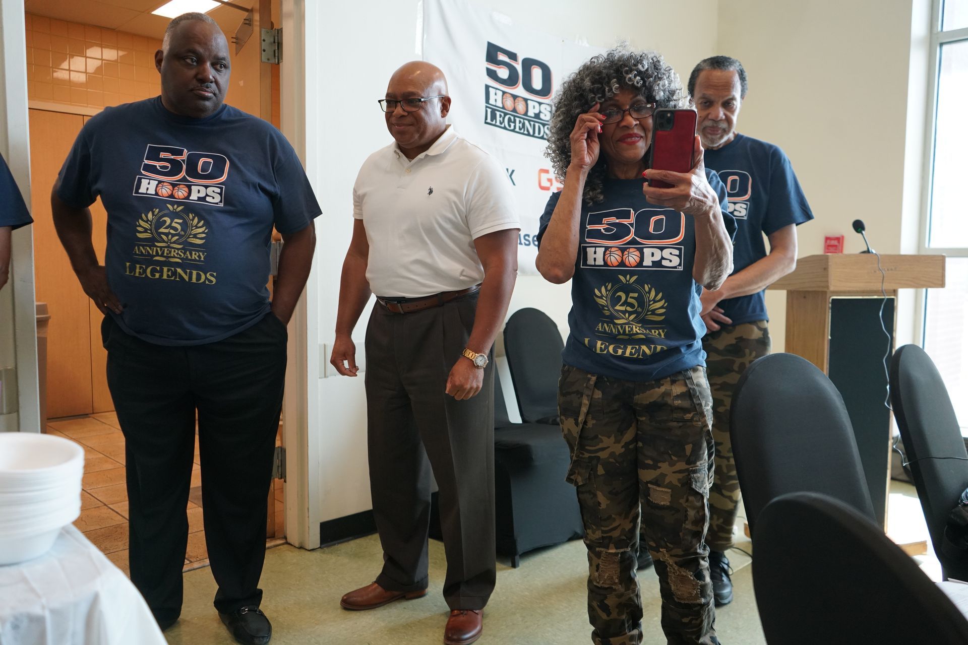 Four people indoors; two in blue shirts with design, others in white shirt and camo pants.