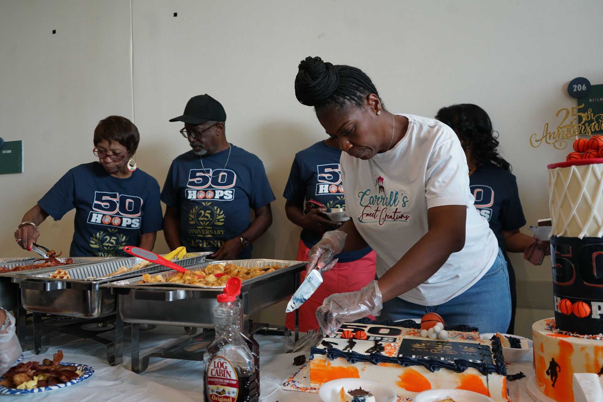 People at a party, serving food from a buffet. A woman cuts cake. Others wear 