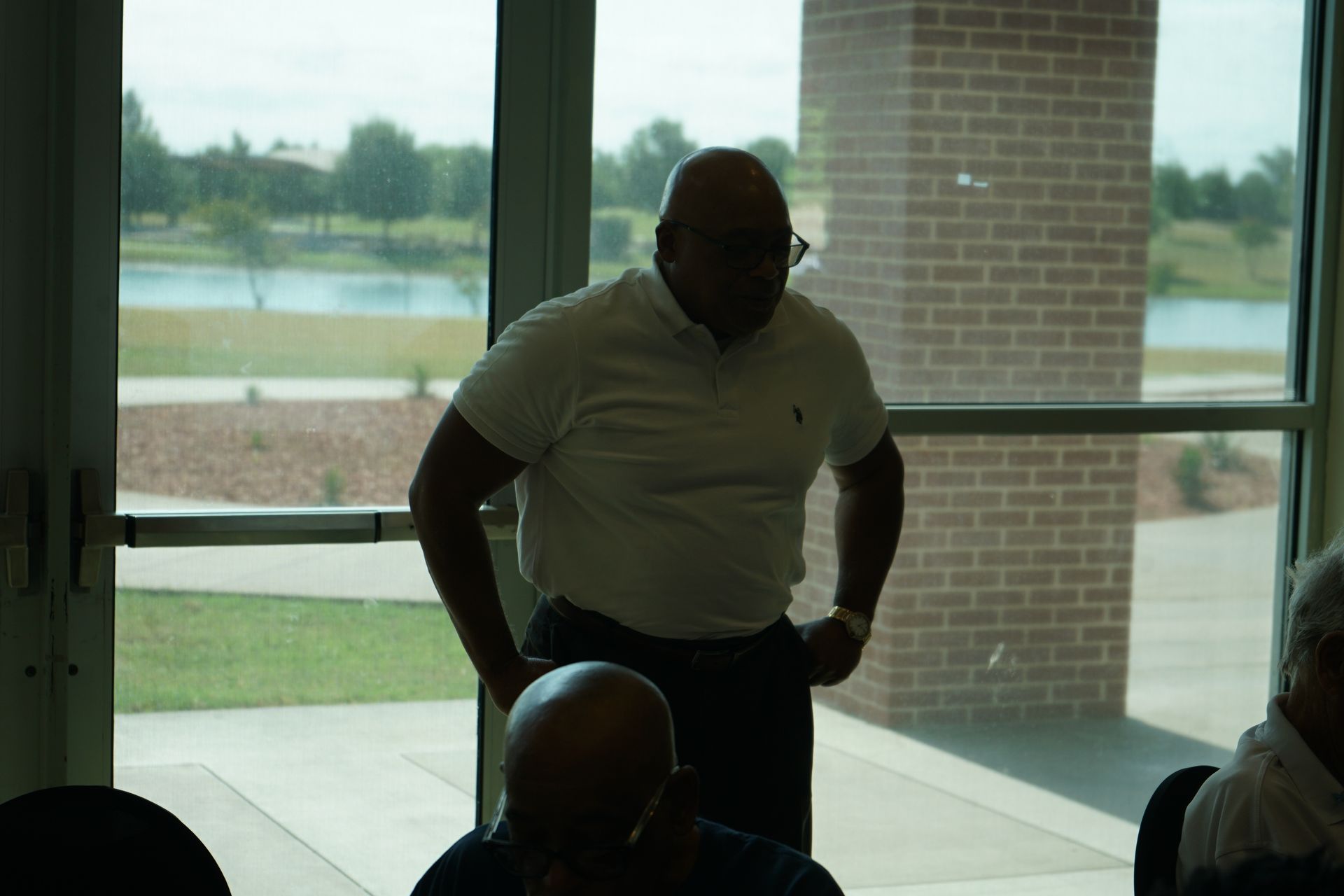 Man in white shirt standing by window, hands on hips, looking down. Brick wall and pond visible.