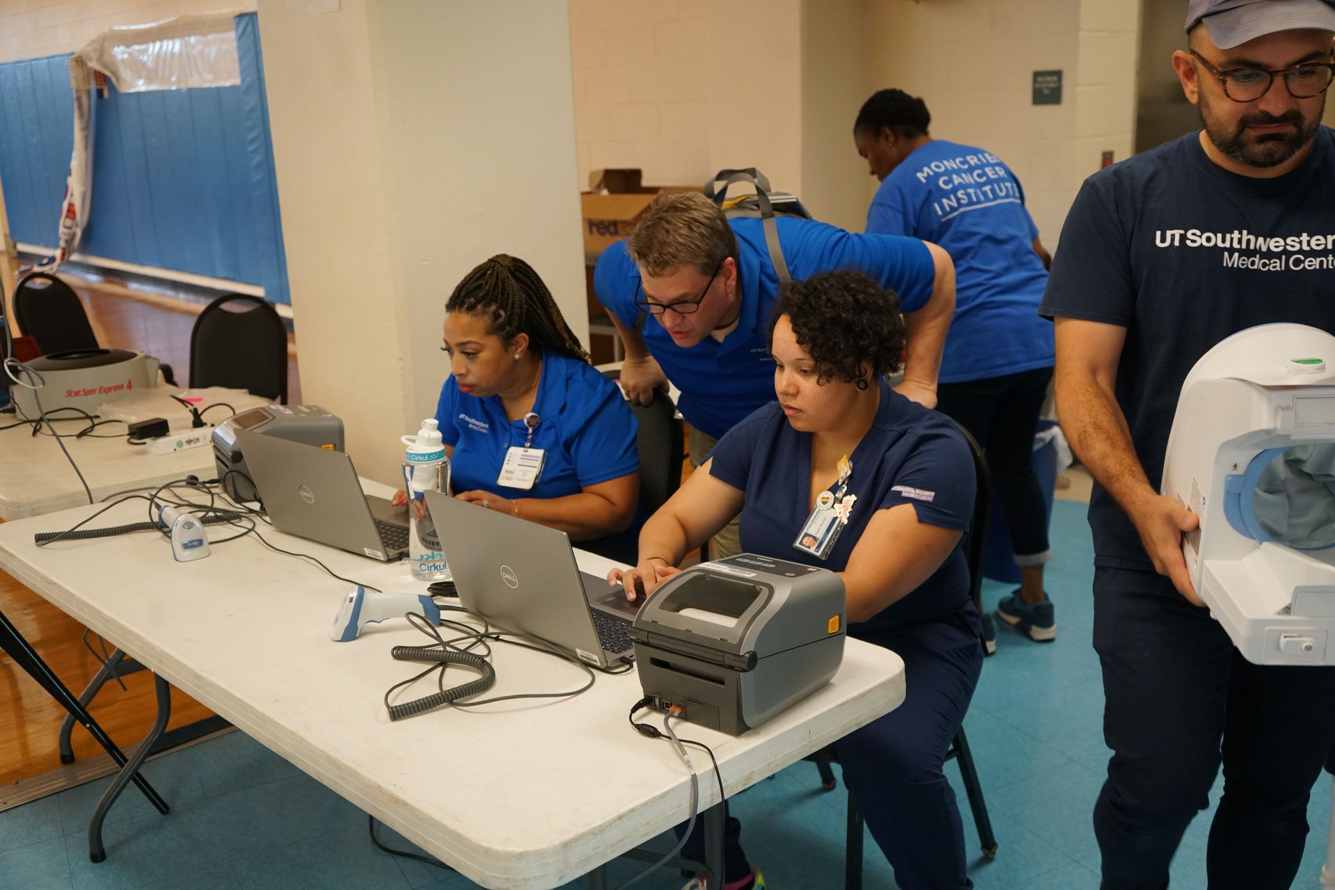 People in blue shirts using laptops and medical equipment at a table. Another person carries a device.