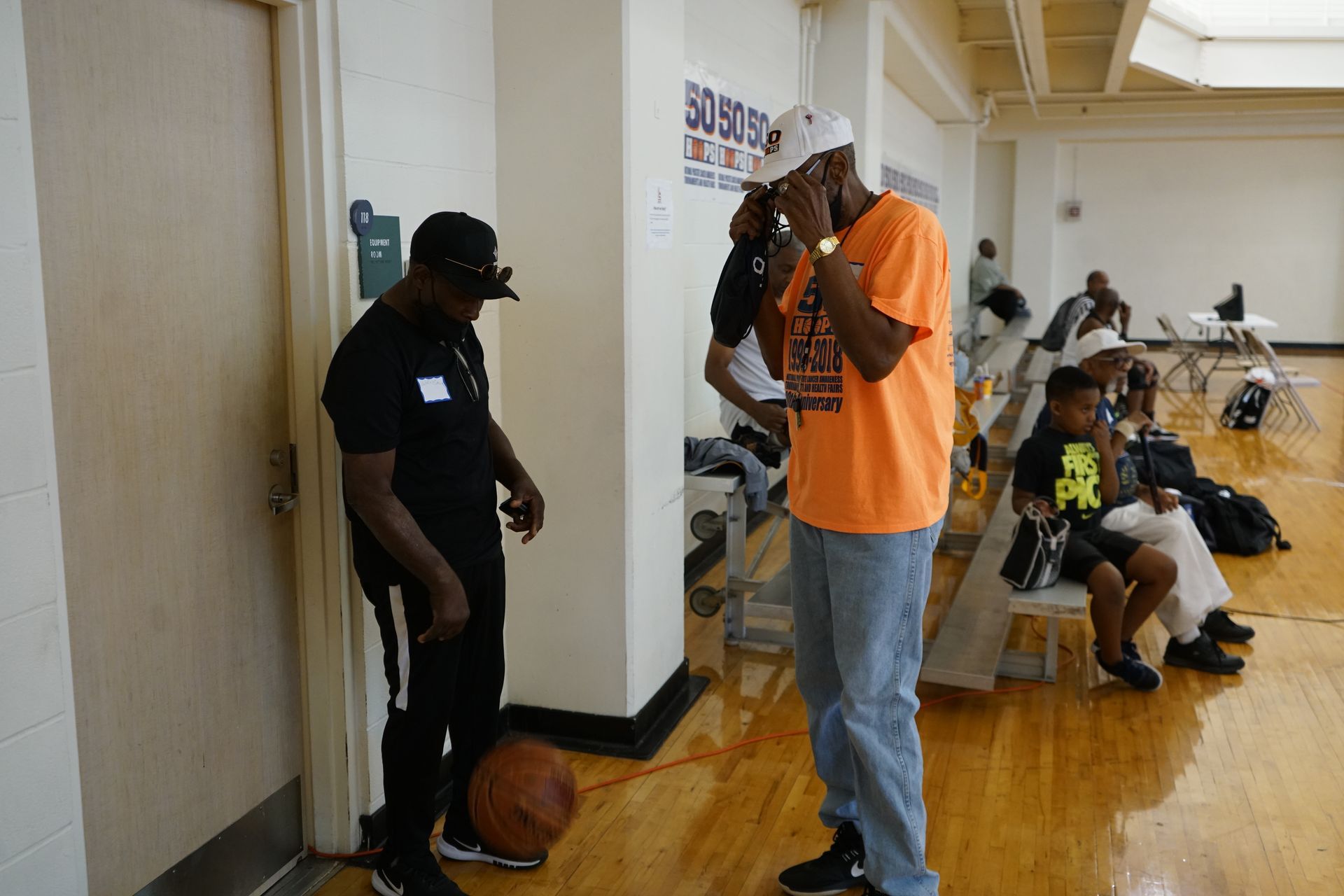 Two men talking near a door inside a gymnasium. One in an orange shirt, the other in black, with a basketball on the floor.