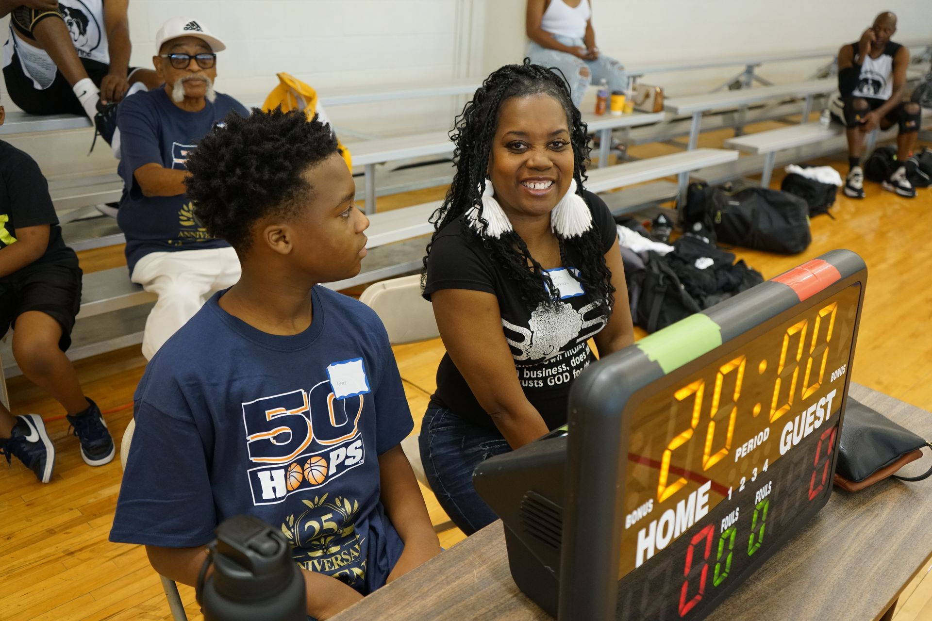 A woman and a young person sit near a basketball scoreboard. Both smile. Spectators watch in the background.