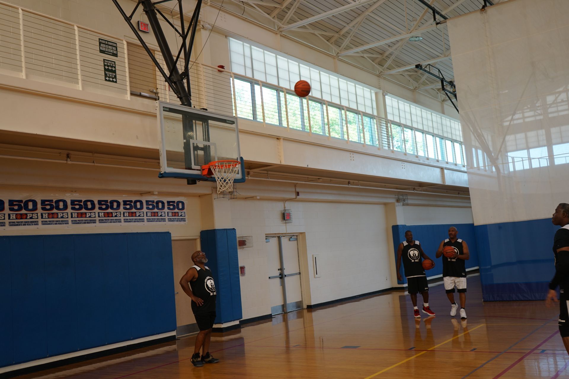Basketball in mid-air, heading for the hoop in a gym. People in black are standing nearby.