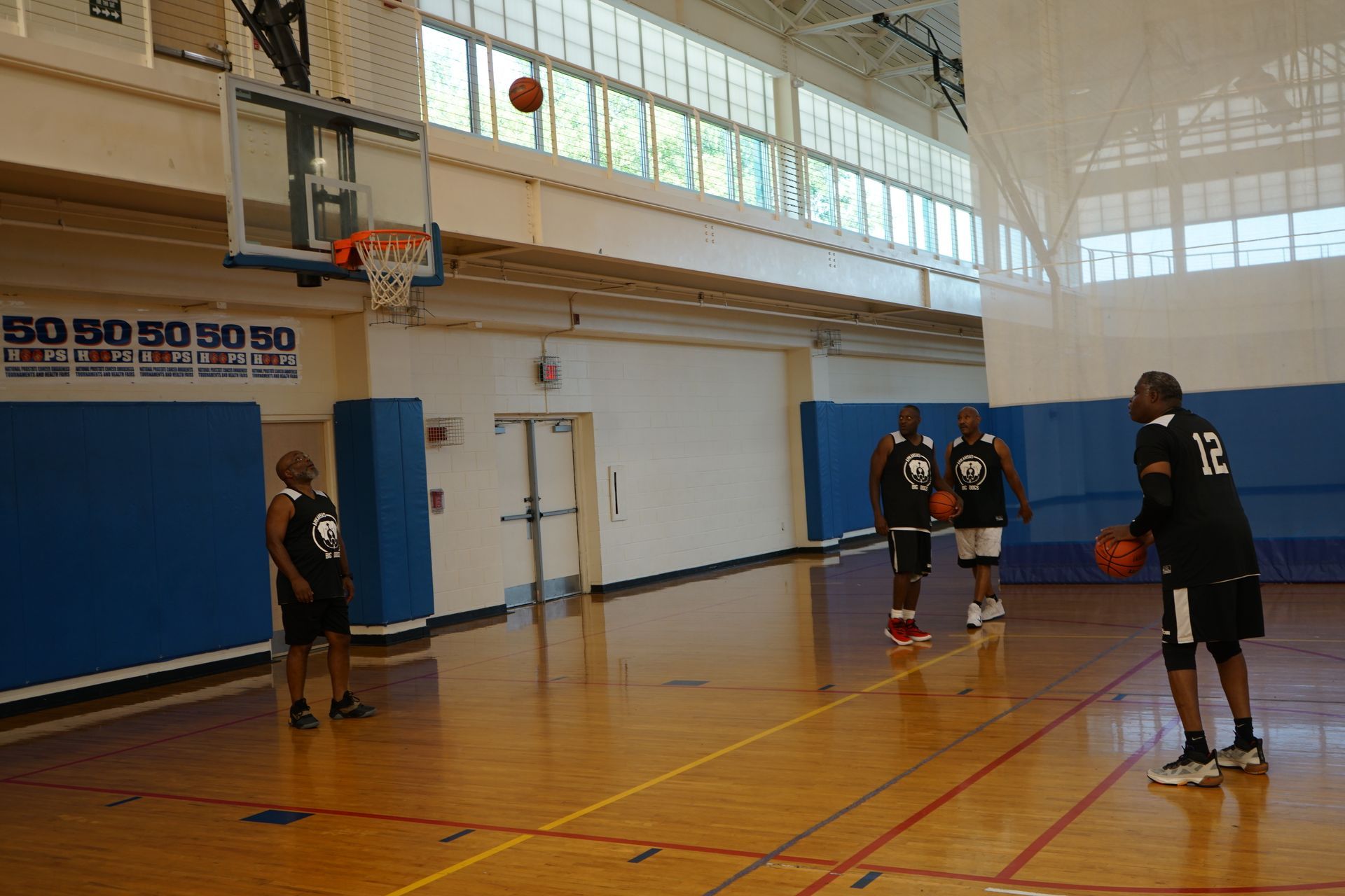 Basketball game in a gymnasium; ball going through the hoop. Two other players watch and one prepares to shoot.