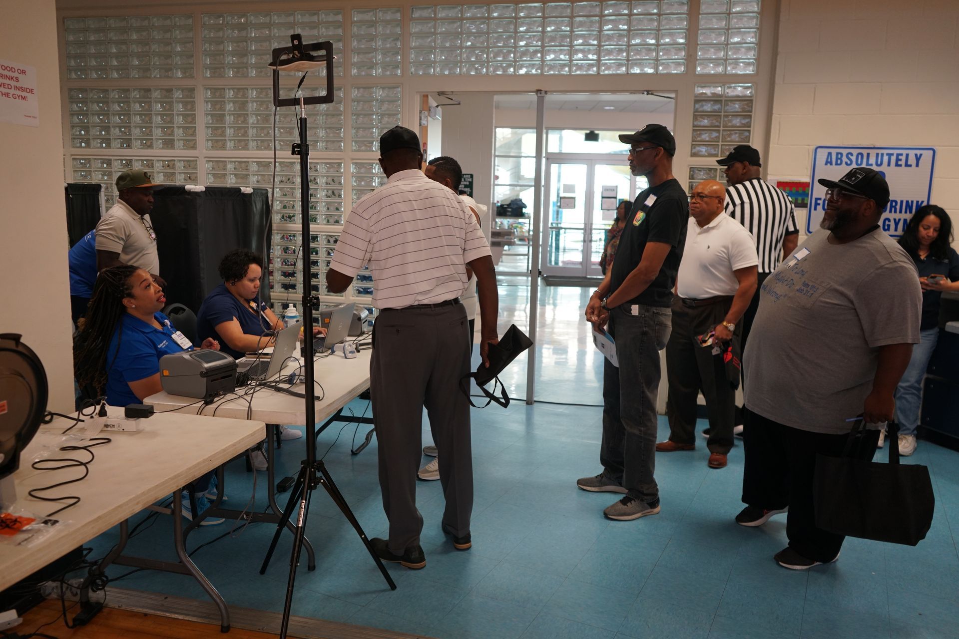 People at a registration or voting site. Some stand in line, others at a table with equipment. Interior setting with a door.