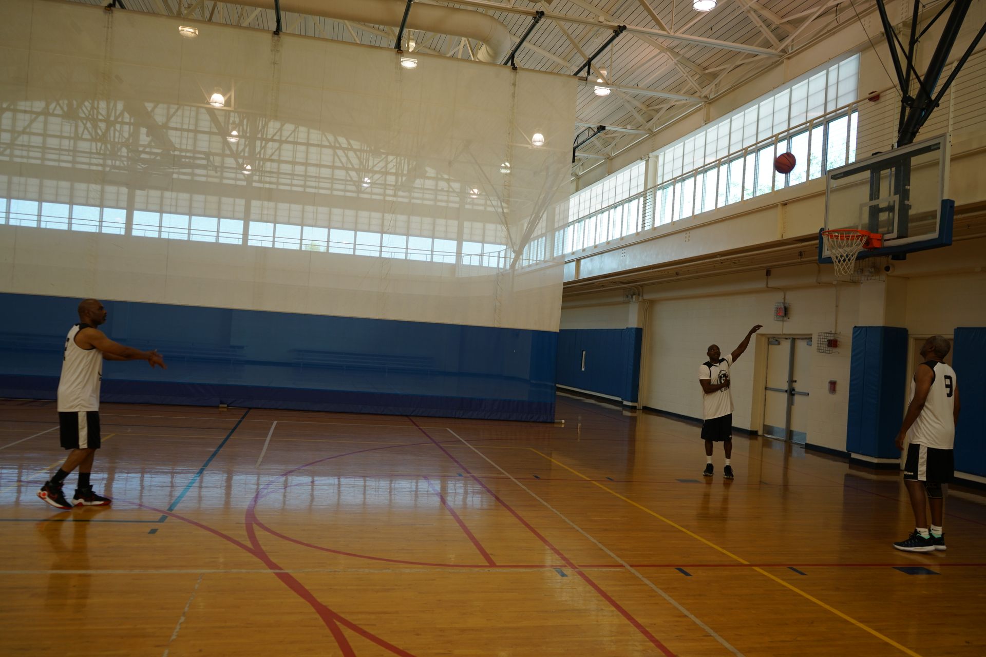Three people playing basketball in a gymnasium. One shoots, two others watch.