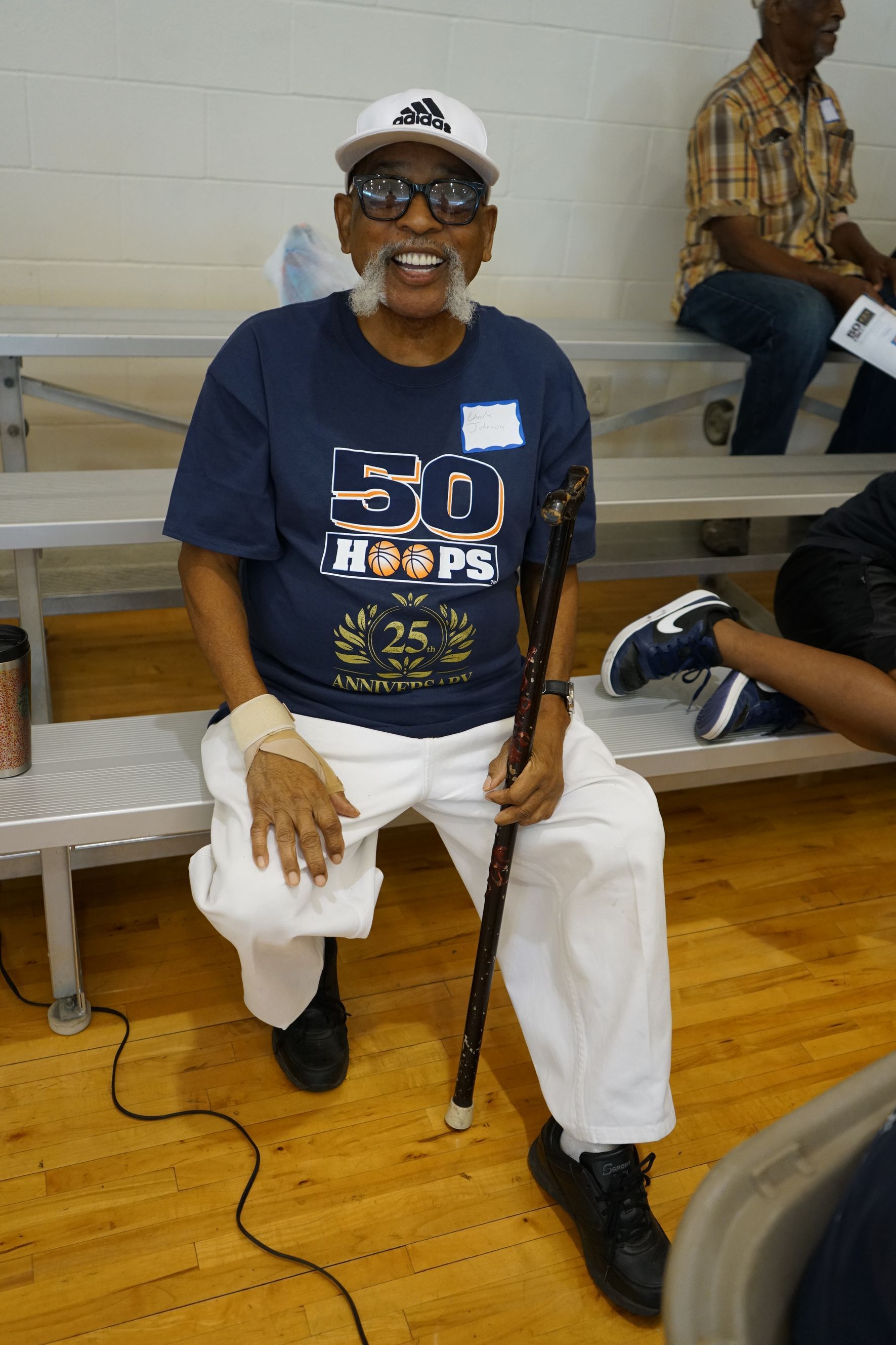 Smiling man wearing a white Adidas cap and blue shirt, sitting with a cane on a bench in a gymnasium.