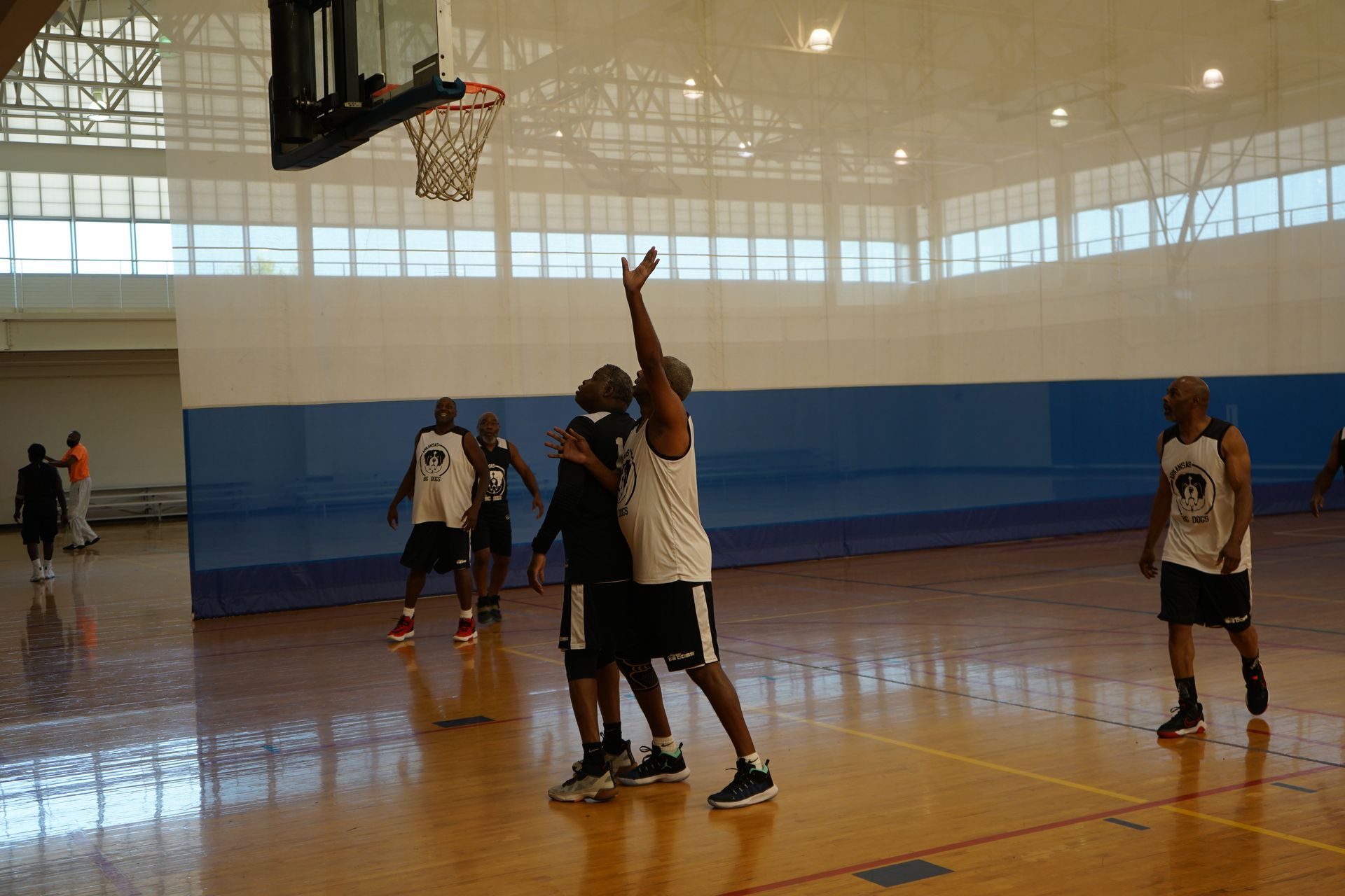 Basketball game in a gym: A player shoots, defended by another. Others watch on a wooden court, blue padding on wall.