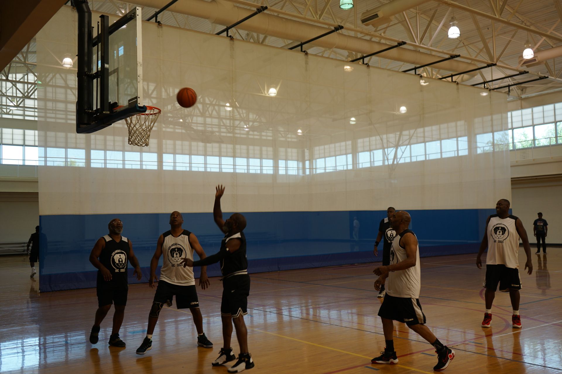 Basketball players in a gym, one shooting towards the hoop. Other players watch.