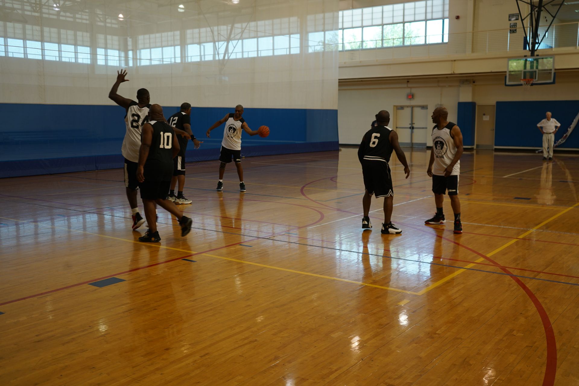 Basketball game in a gym. Players in jerseys dribble and pass. Wooden floor, white walls, and blue accents.