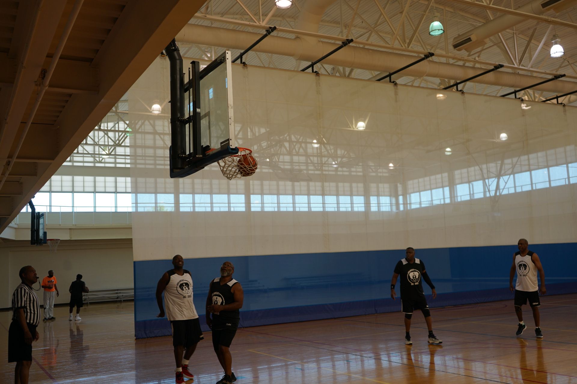 Basketball game in a gym: ball going into hoop, players on court, some wearing jerseys, blue and white walls.