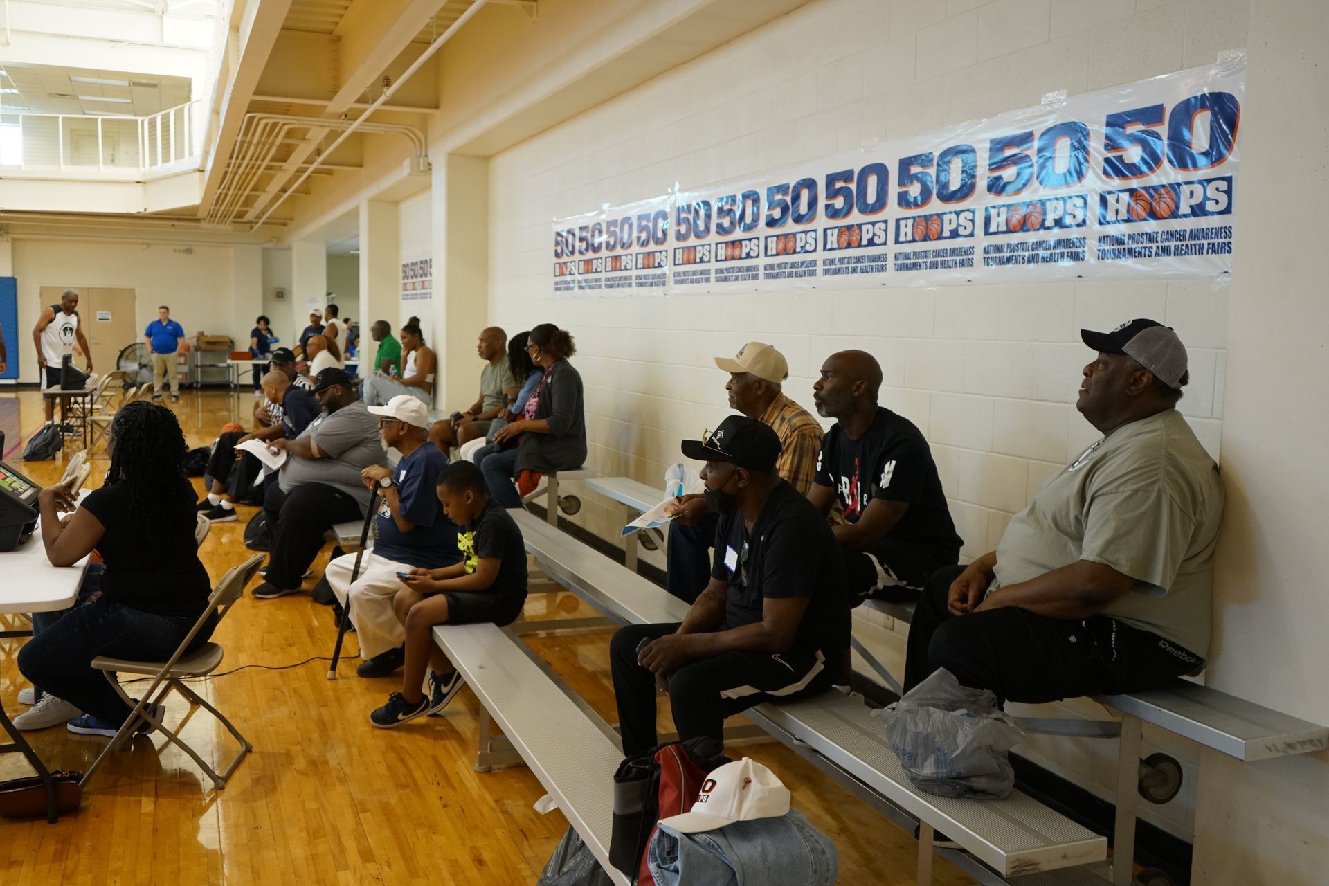 People seated in bleachers watching something in a light-filled gym with a banner that reads 