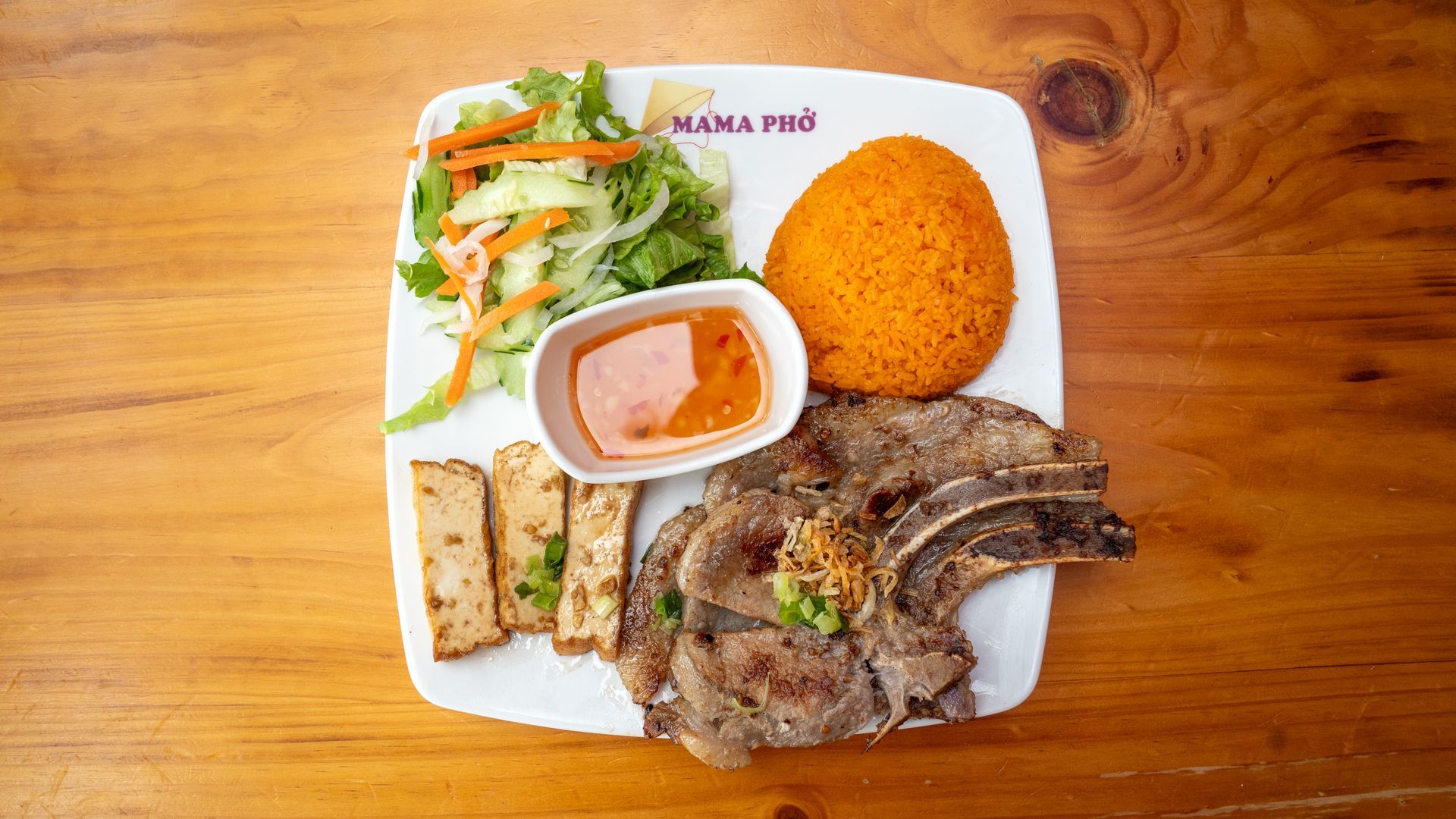 A white plate topped with meat , rice , and vegetables on a wooden table.