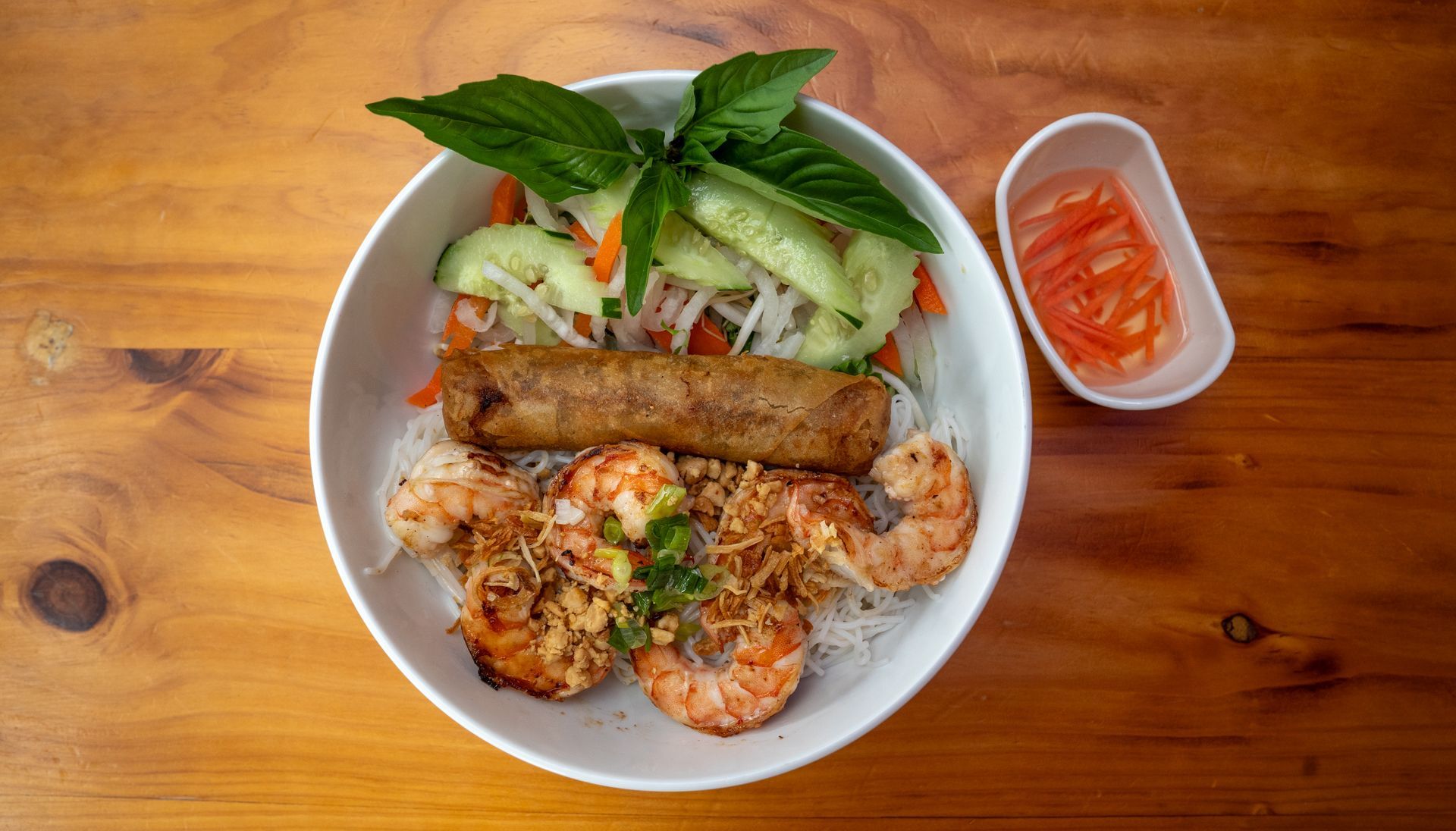 A bowl of food with shrimp , spring rolls and vegetables on a wooden table.