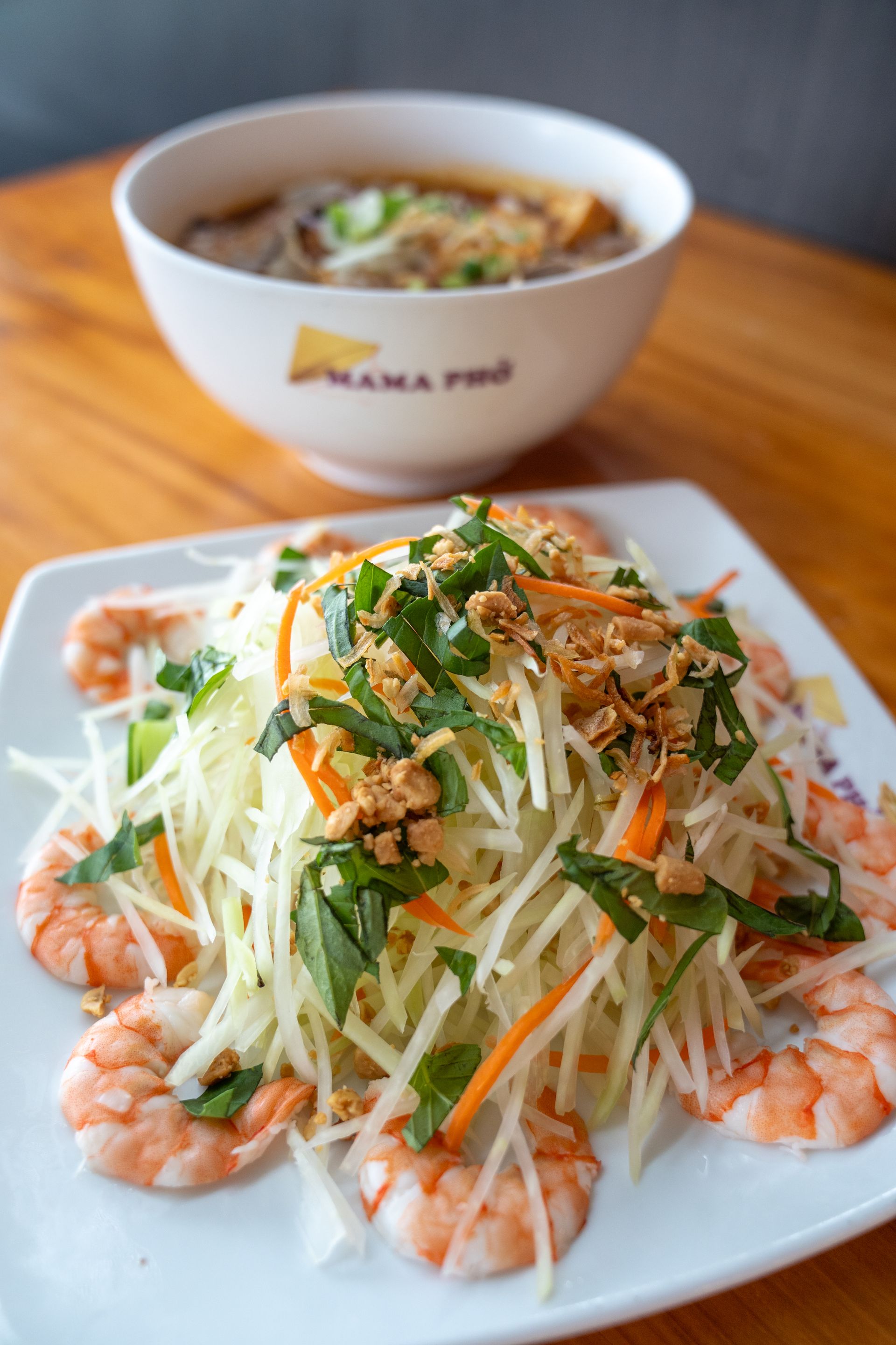 A plate of food with shrimp and vegetables next to a bowl of soup on a table.