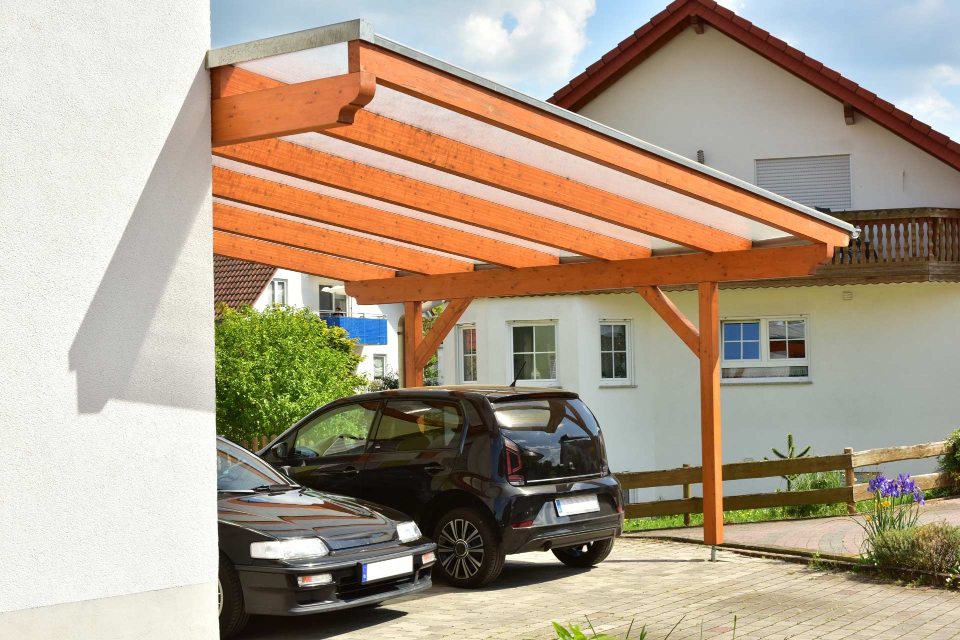 Two cars are parked under a wooden carport in front of a house