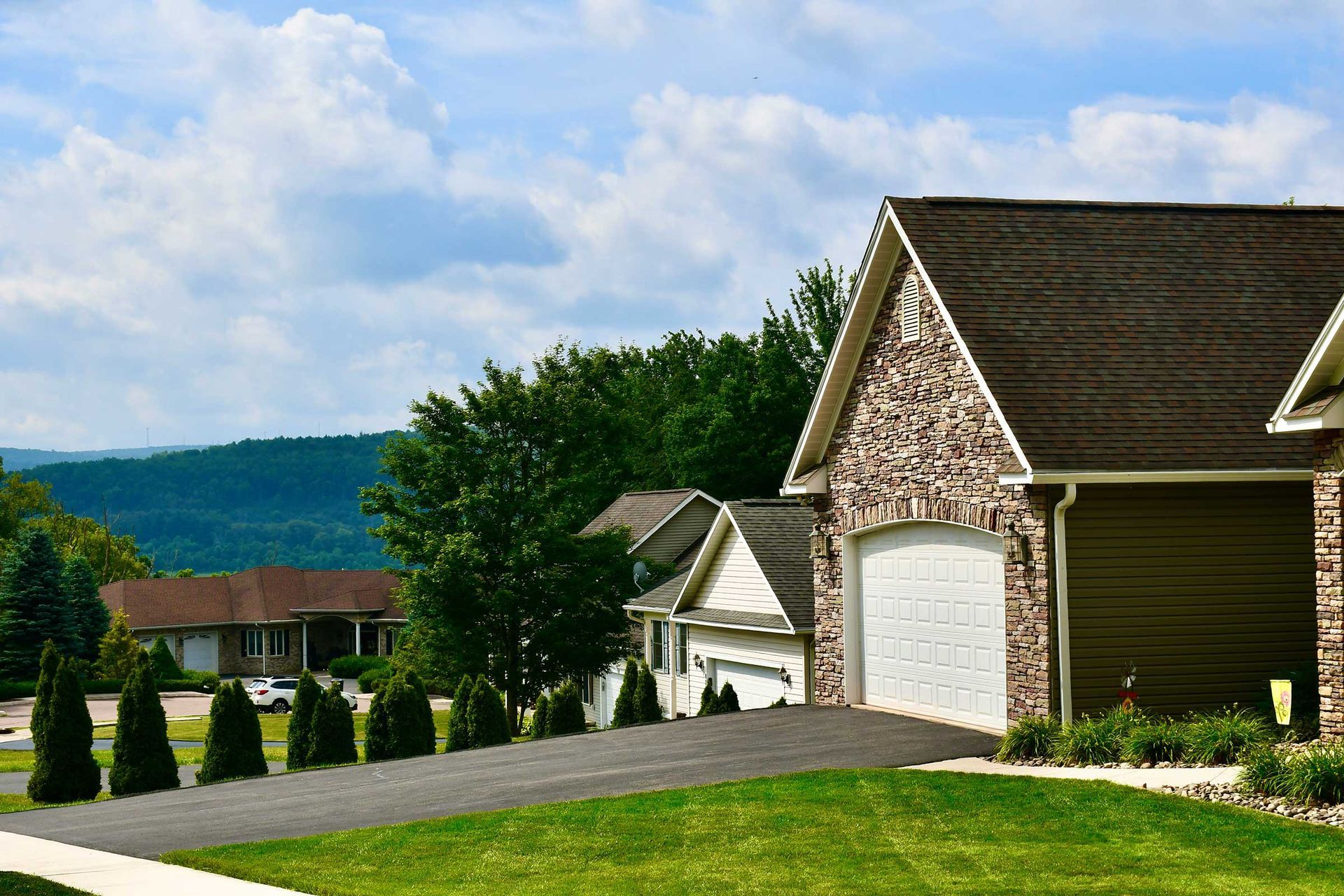 Houses with driveways and manicured lawns on a hillside under a blue sky with clouds; trees and mountains are in the background.