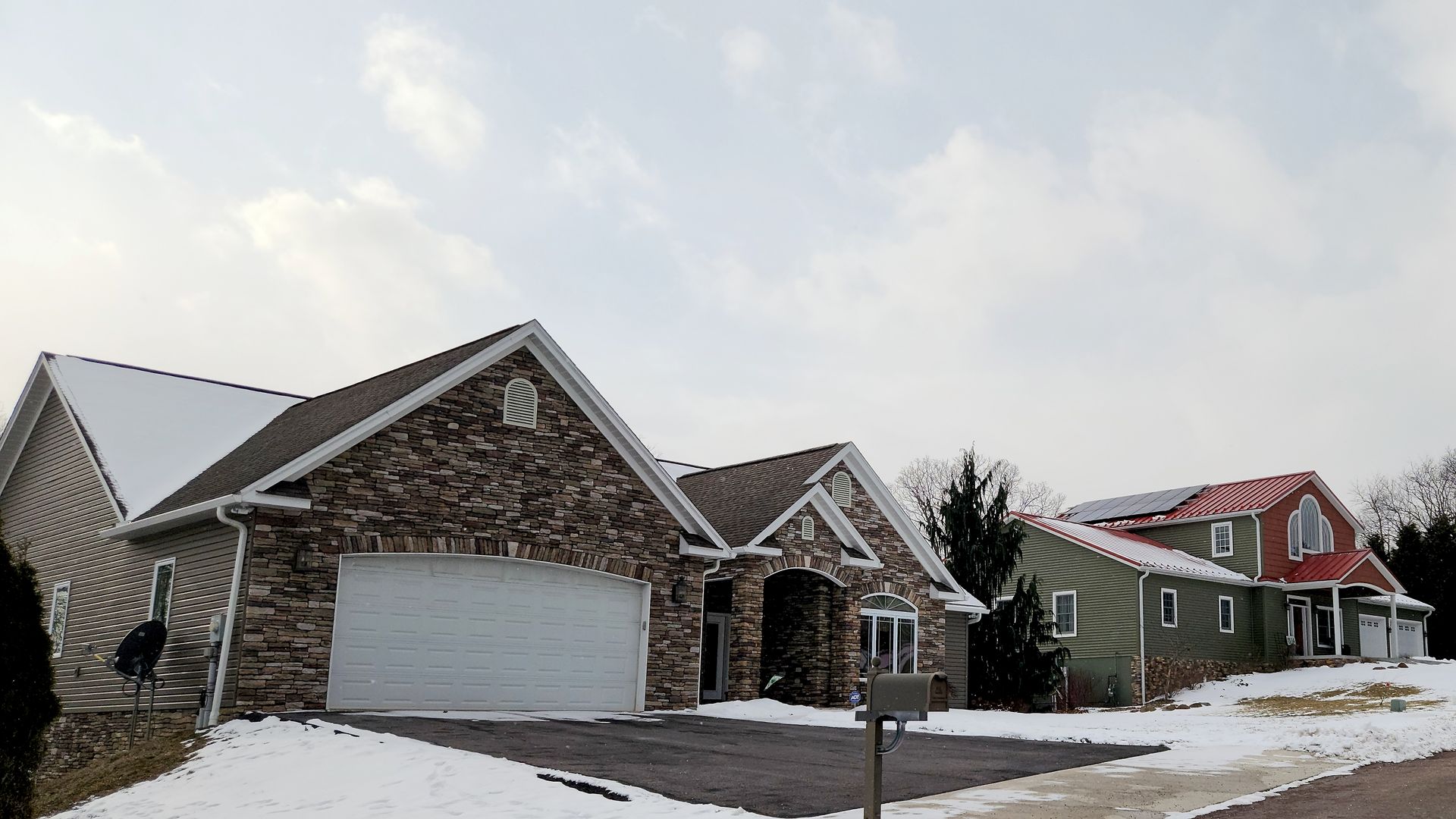 Houses covered in snow on a cloudy day. One house has a stone facade and attached garage. Another is green with a red roof.