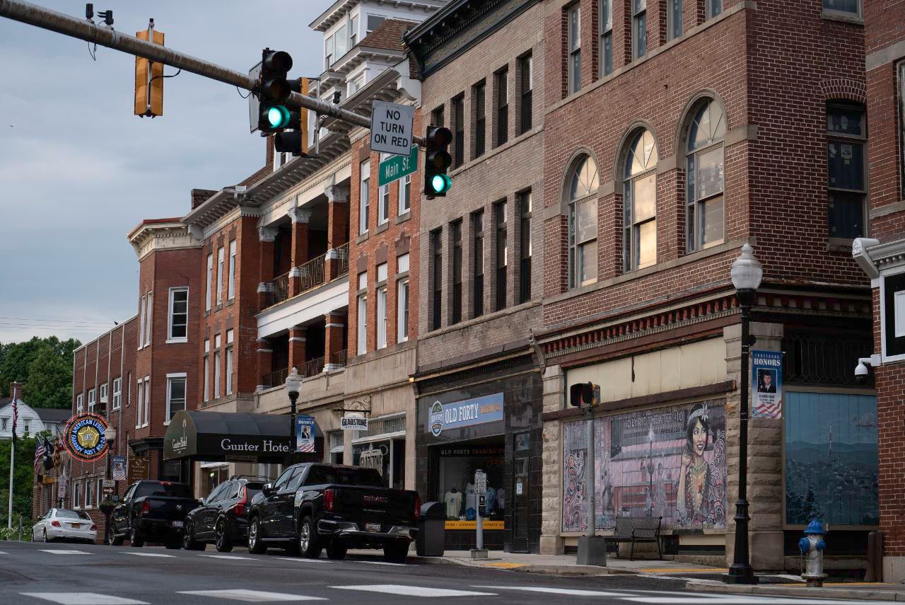 Street scene with brick buildings, parked cars, and traffic lights. Green traffic lights are visible.